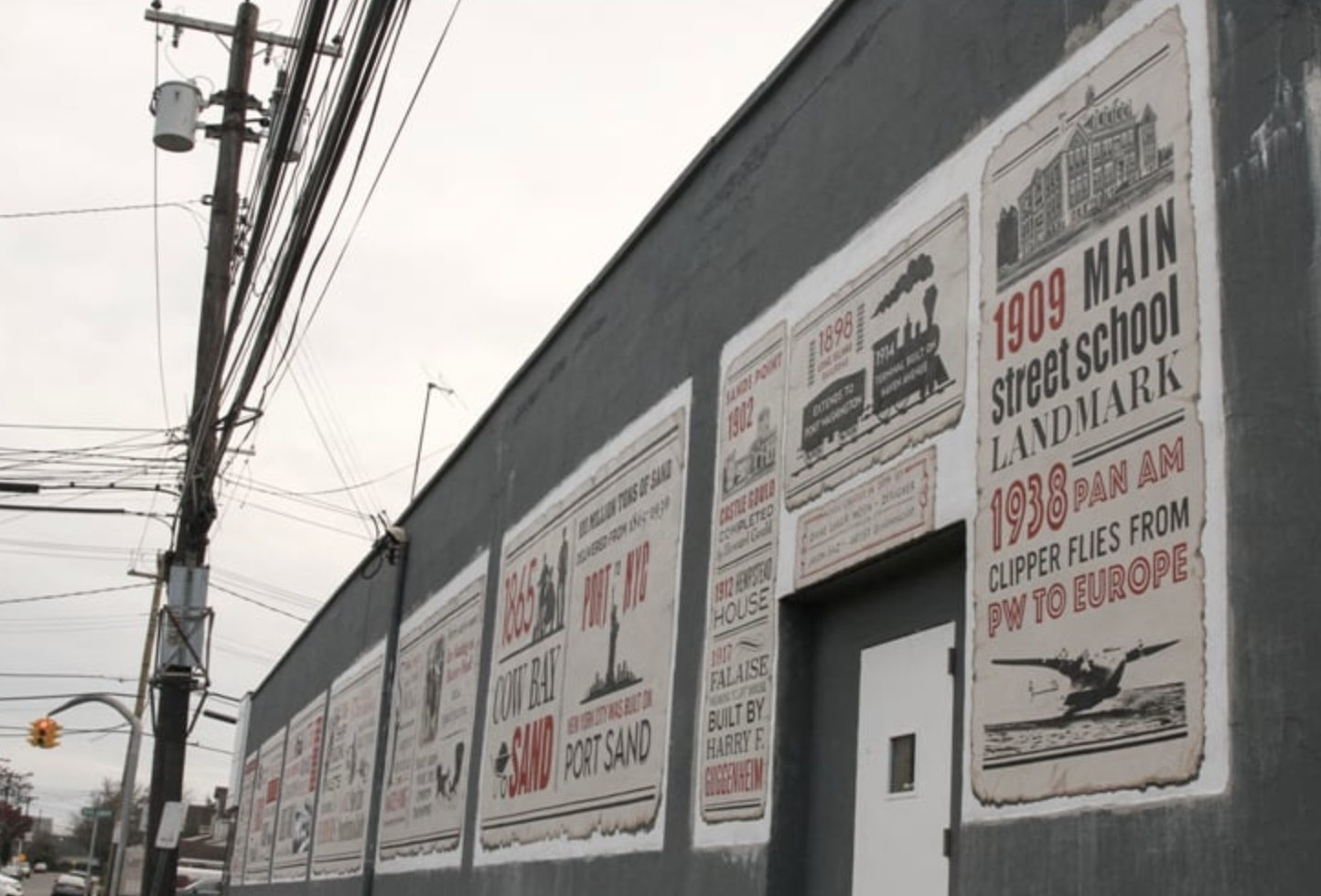 Historical murals on a building wall featuring vintage posters about local landmarks and past events, with power lines and a traffic light in the background.