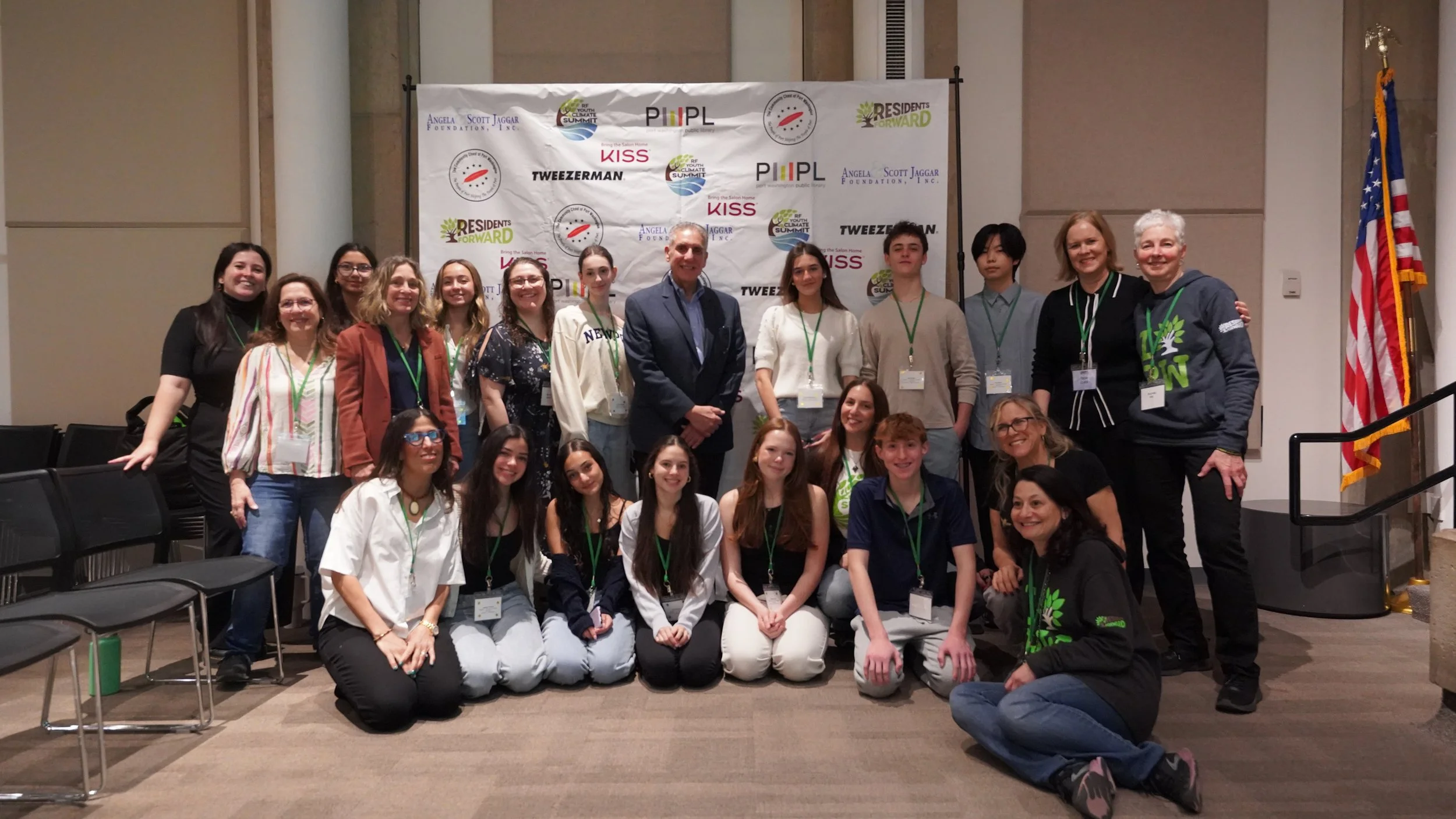 Group of people posing for a photo in front of a backdrop with various logos, including Angela Scott Jaggar Foundation, RESIDENTS FORWARD, KISS, and others, at an indoor conference or event. The group includes both adults and young people, some wearing name tags and green lanyards.