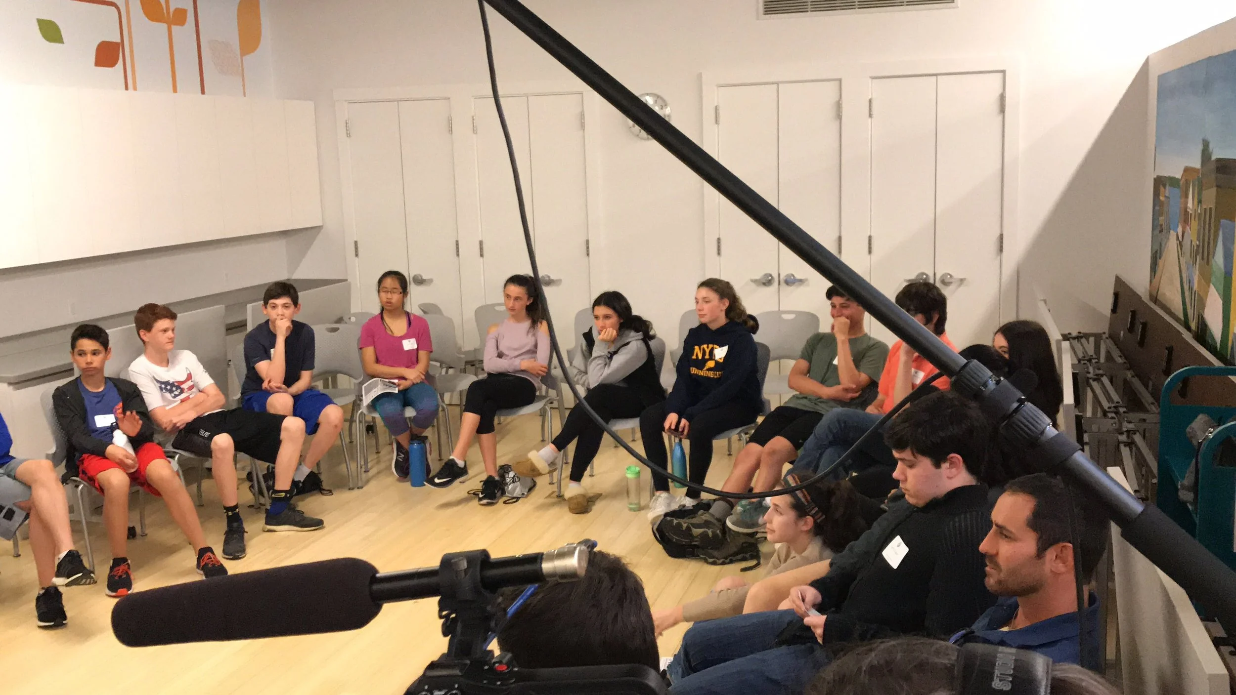 Group of children and teenagers seated in a circle in a room, engaged in activity or discussion, with a camera and microphone visible in the foreground.