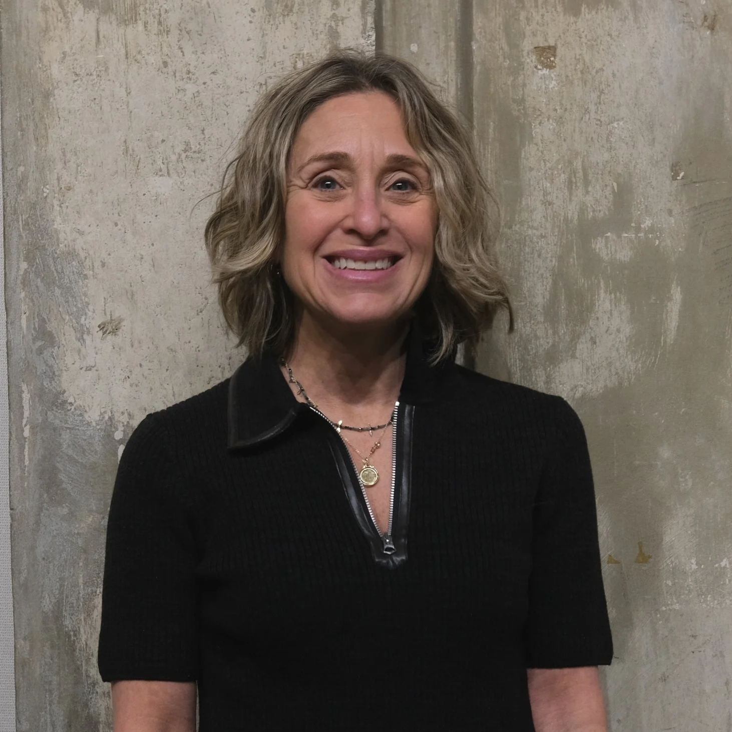 A woman with shoulder-length wavy blond hair, smiling, wearing a black top with a zipper neck, a gold necklace, and standing against a textured concrete wall.