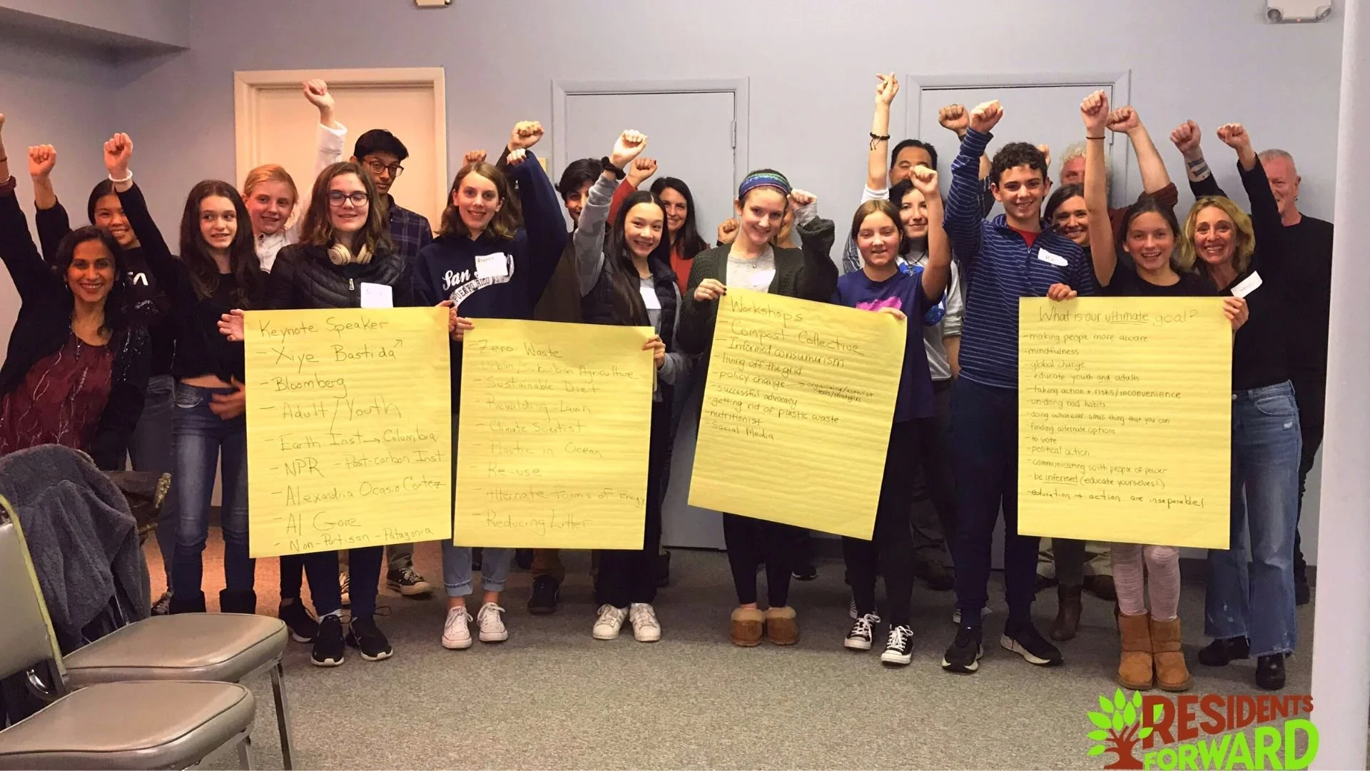 Group of young people and adults standing indoors, holding large yellow posters with handwritten notes, raising fists in solidarity, smiling, in a room with light blue walls. Some chairs are visible in the foreground, and a logo for "Residents Forward" is on the bottom right corner.