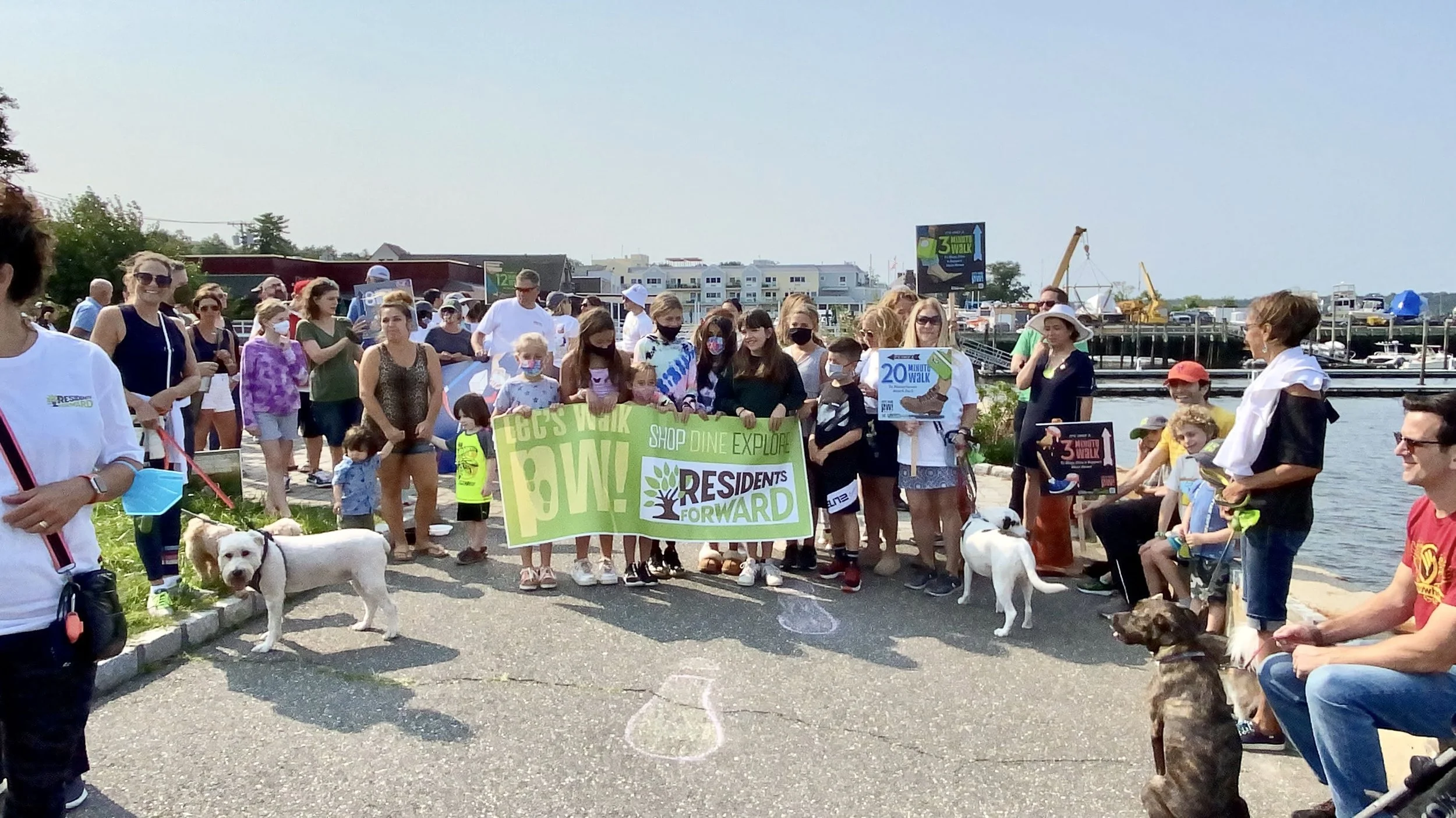 A large group of people, including children and adults, gathered near a waterfront for a community event. Some are holding signs that say "Let's Walk 3 Miles," "Residents Forward," and related messages, with dogs accompanying them.