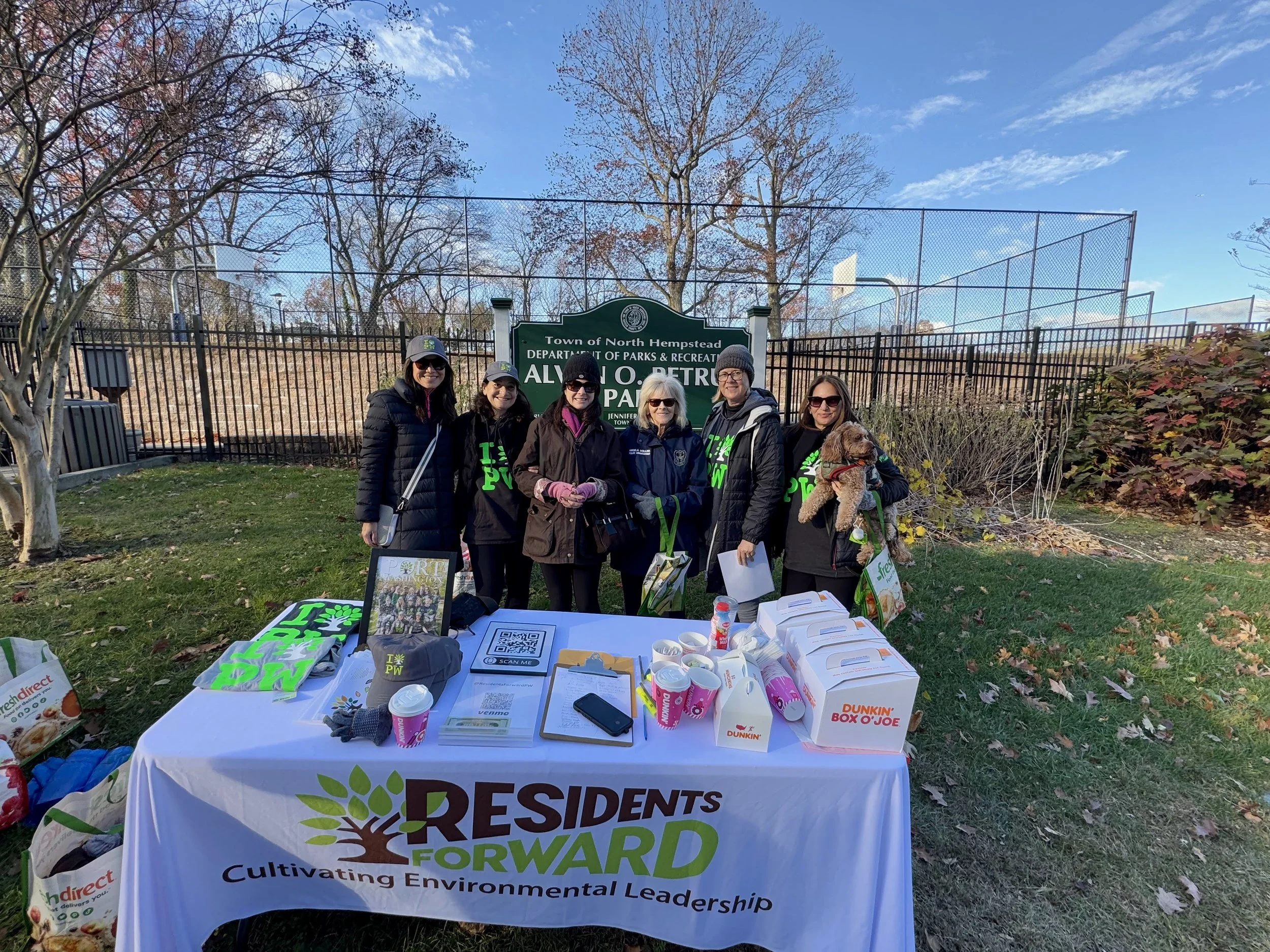 Group of six women standing behind a table with community resources at a park in North Hempstead, with a sign for Alvano O. Petruccelli Park in the background.