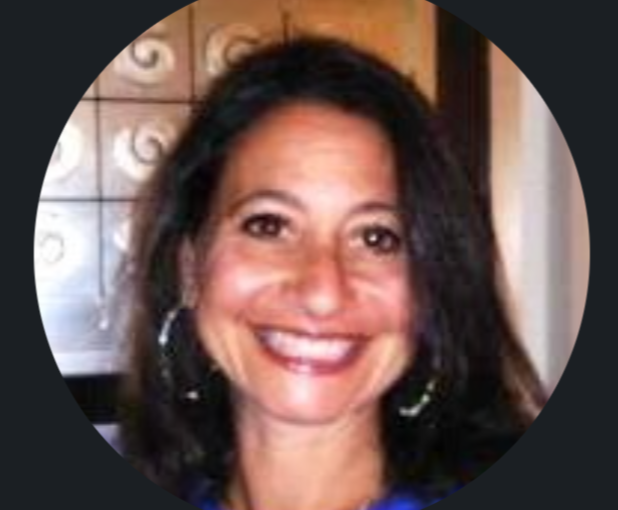 A woman with long dark hair smiling, wearing hoop earrings, in front of a wooden background.