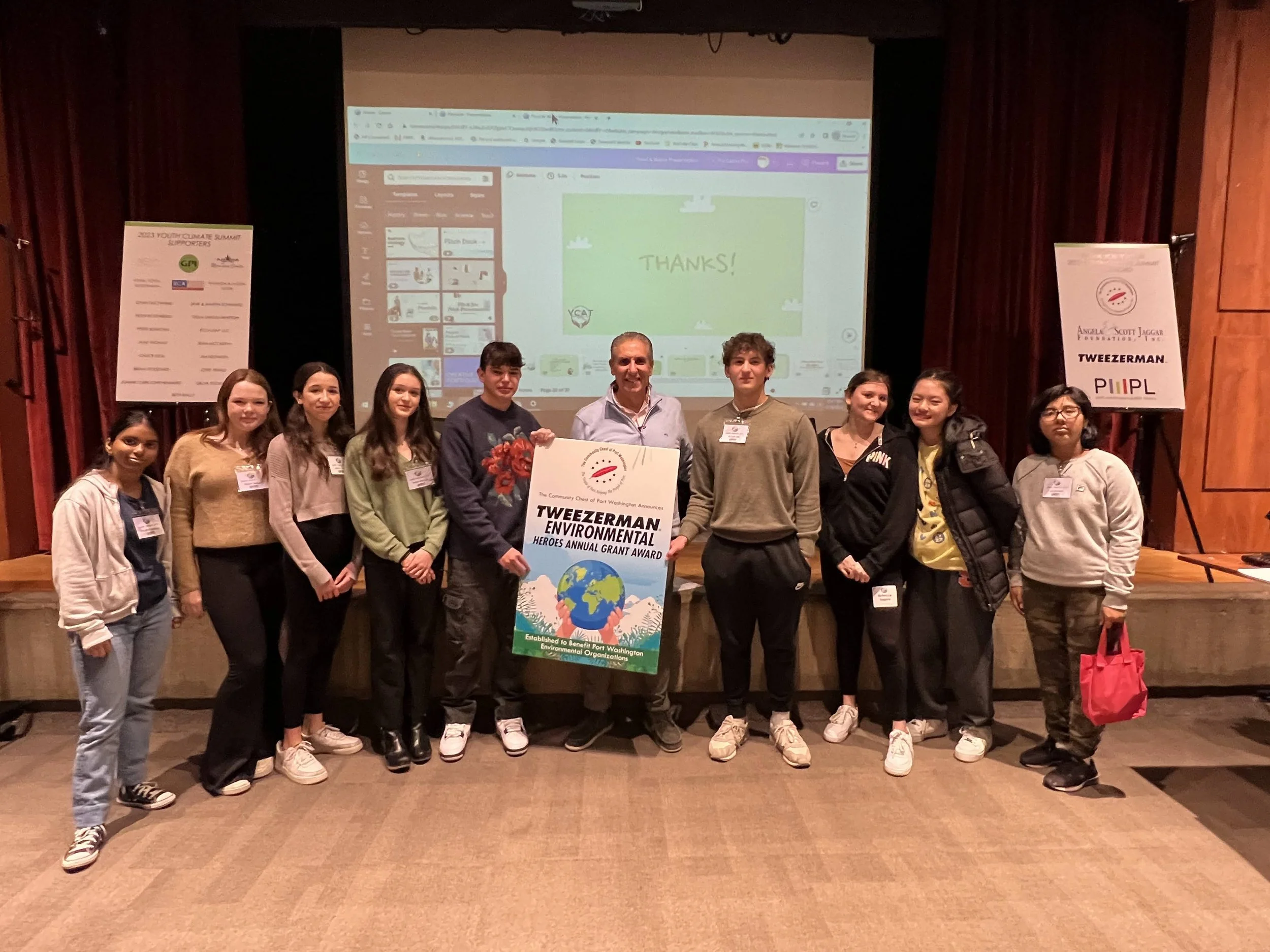 Group of young students and an adult on stage holding a large award sign that says "Tweezerman Environmental Heroes Annual Grant Award" at an event, with a presentation screen in the background displaying a "Thanks!" message.