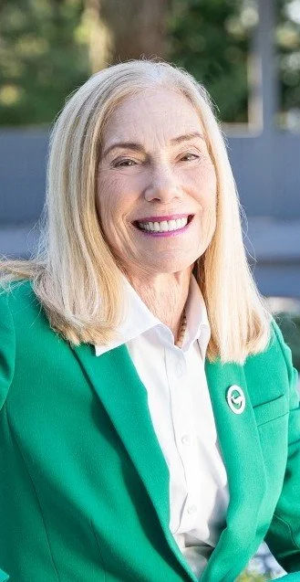 A smiling middle-aged woman with blonde hair, wearing a white shirt and green blazer, outdoors with trees and a fence in the background.