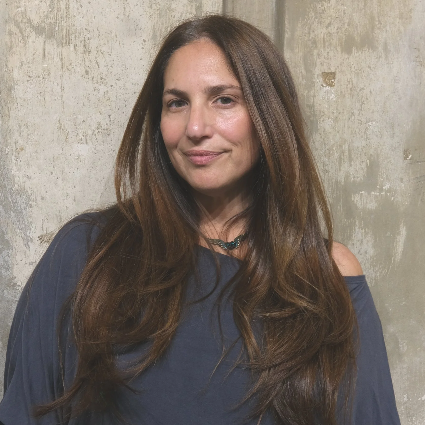 A woman with long, wavy brown hair wearing a dark blue off-shoulder top and a dark necklace, standing against a textured beige wall.
