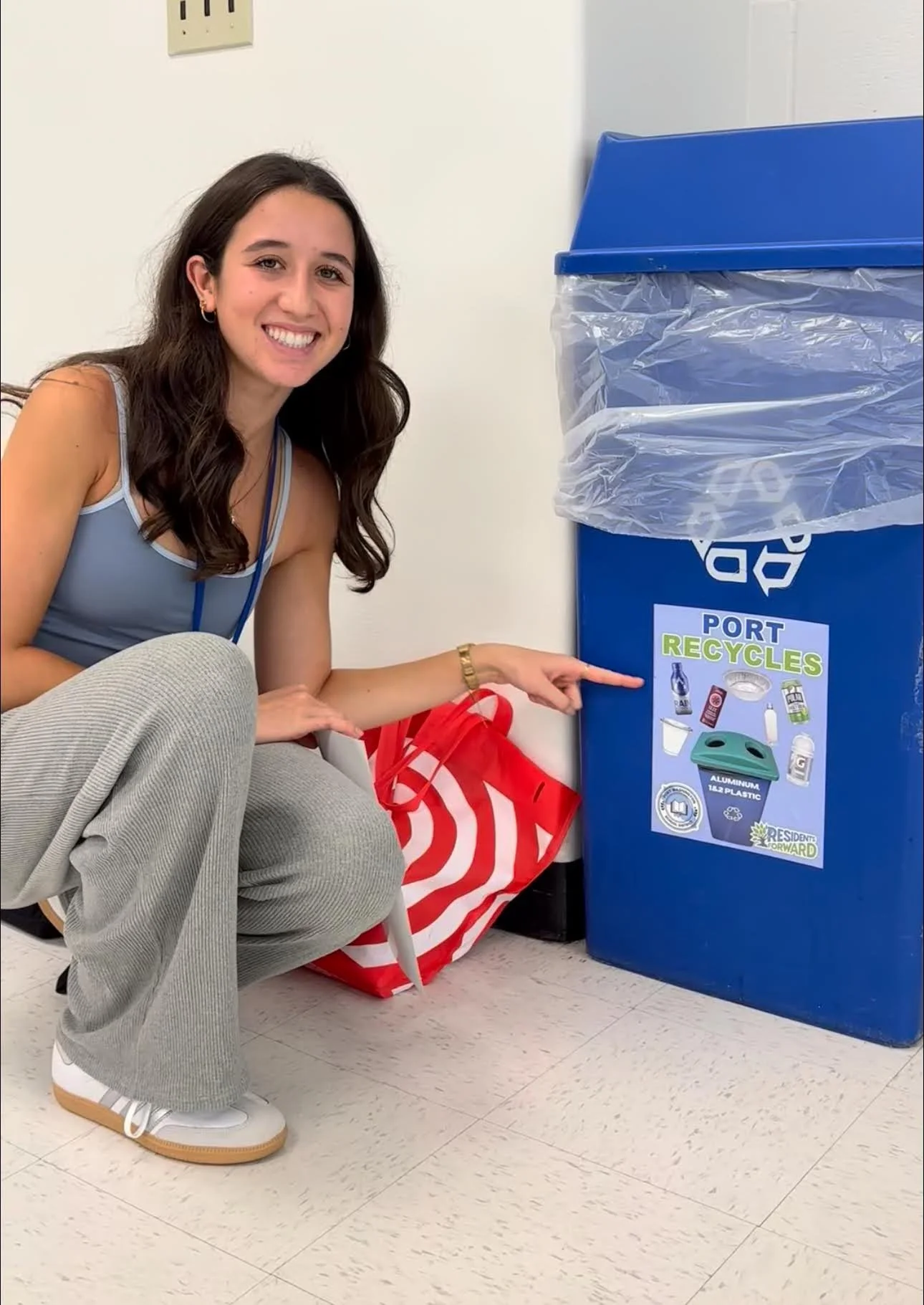 A young woman with long dark hair squatting next to a blue recycling bin, pointing at a sign on the bin that says 'Port Recycles' with images of recyclable items. She is smiling and wearing a sleeveless top, gray pants, white sneakers, and a watch, w