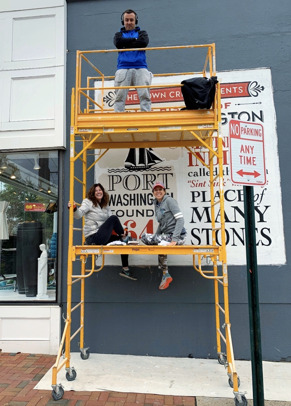Two women sit on a yellow scaffold in front of a mural, one on the lower level and one on the upper level, smiling and posing for the photo.