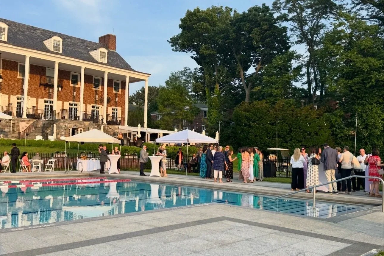 Guests gather by a swimming pool at the Residents Forward Gala - near a large brick mansion the Manhasset Bay Yacht Club, surrounded by trees and greenery.