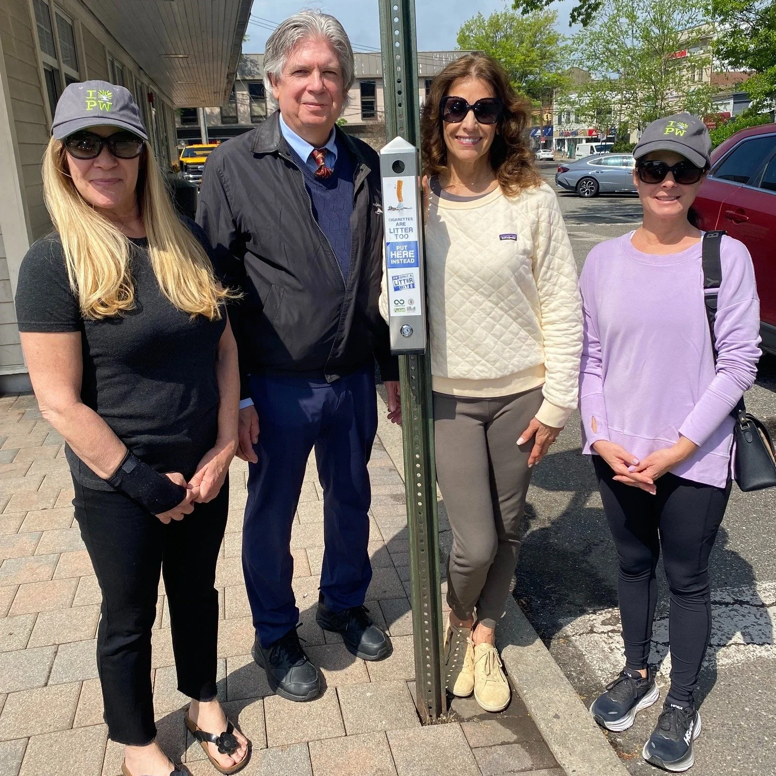 Four women and one man standing outdoors on a sidewalk next to a parking meter, with cars and buildings in the background. Two women are wearing Washington State Parks hats.