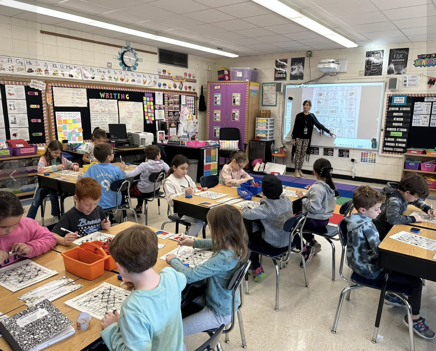 Classroom with young students seated at tables working on worksheets, a teacher at the whiteboard, colorful decorations, and educational materials on the walls.
