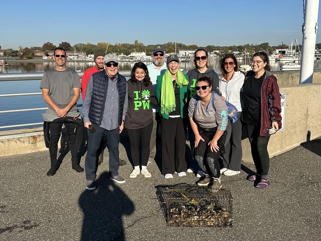 Group of people posing outdoors near a body of water with boats, some wearing sunglasses and outdoor clothing, standing behind a cage filled with discarded vegetables or shellfish.