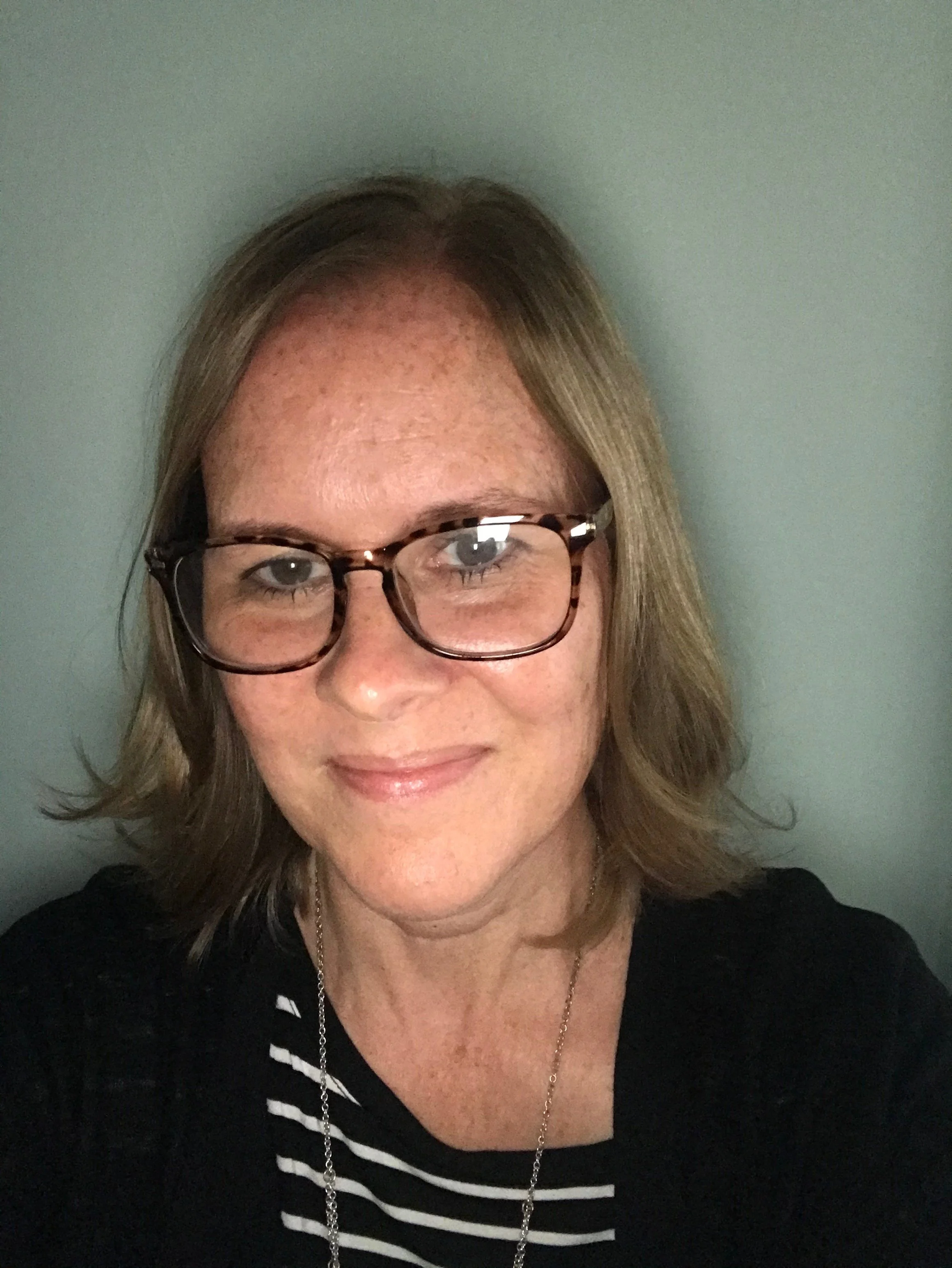 Patricia Class, A woman with glasses and shoulder-length hair smiling at the camera, wearing a black and white striped shirt and a silver necklace.