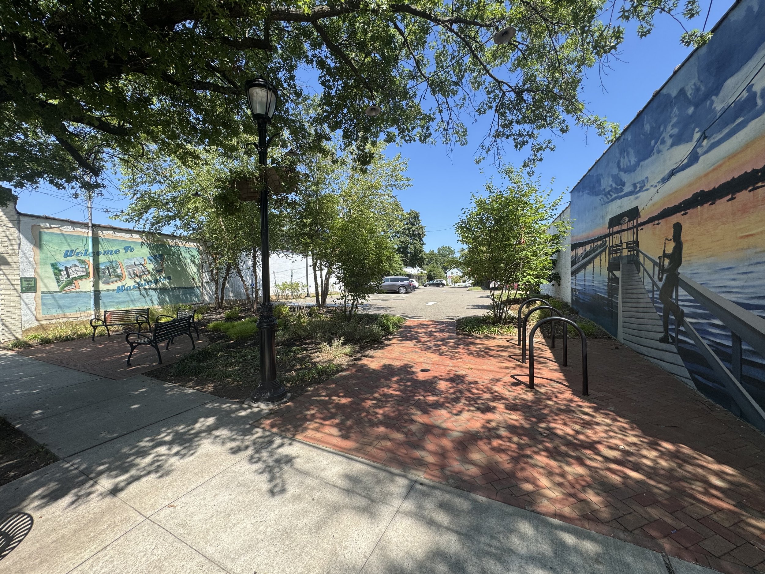 A small outdoor area with benches, trees, a lamppost, and murals on the walls depicting a sunset at the pier, with a parking lot in the distance.