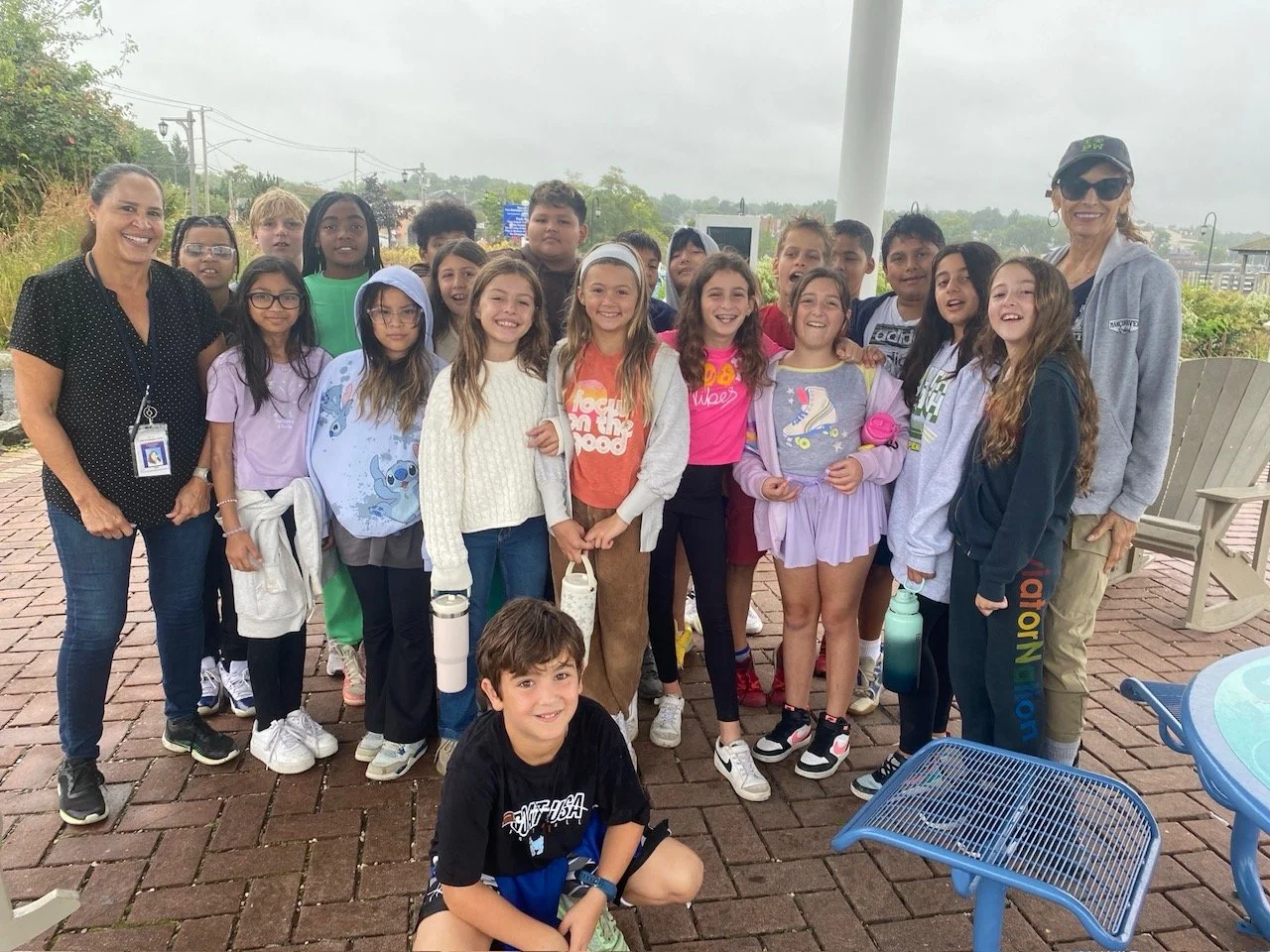 Group of children and two adults smiling outdoors on a rainy day, standing on brick paving with some outdoor furniture around.