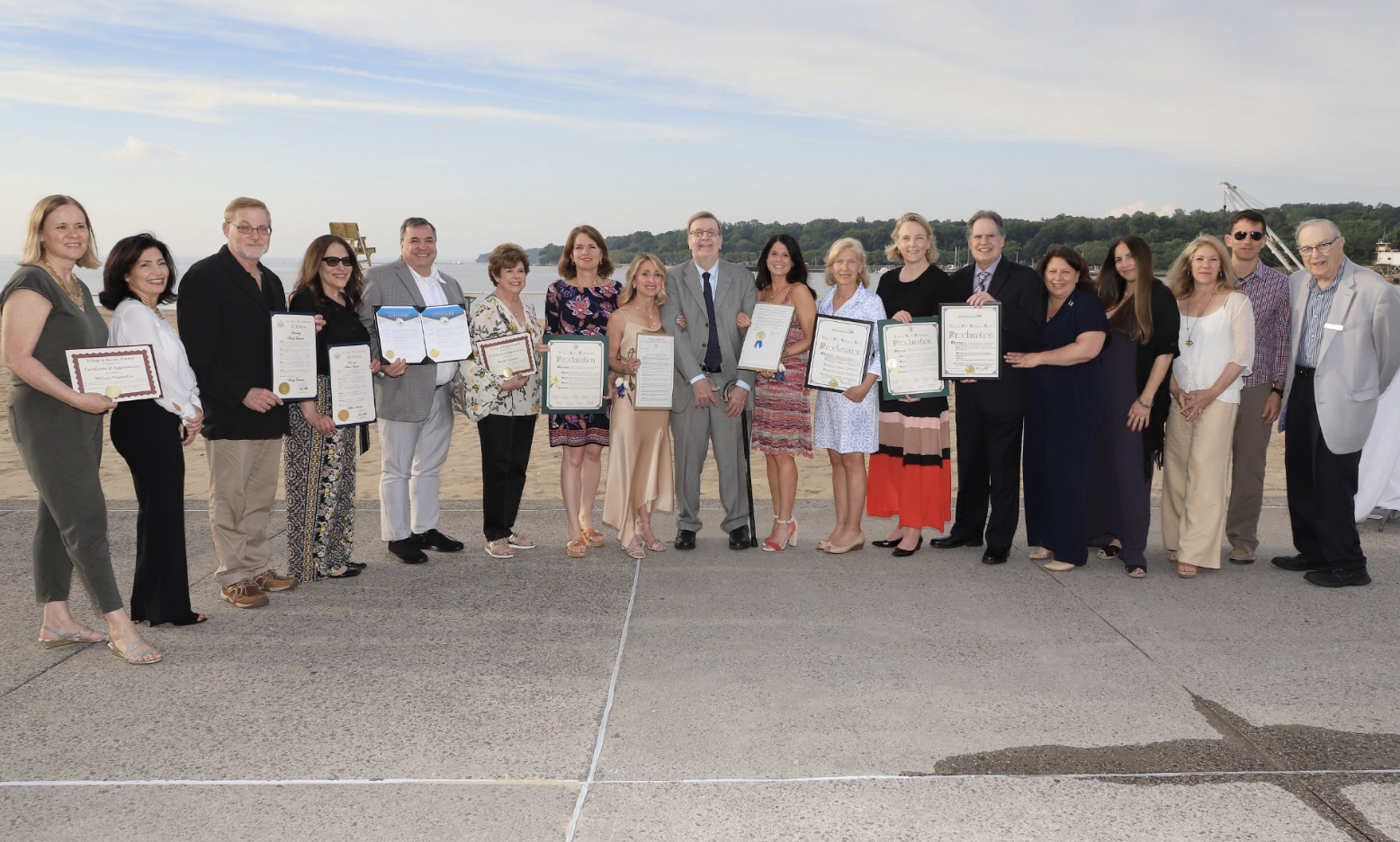 Group of people standing outdoors on a beach, holding certificates, with water and a crane in the background, during sunset.