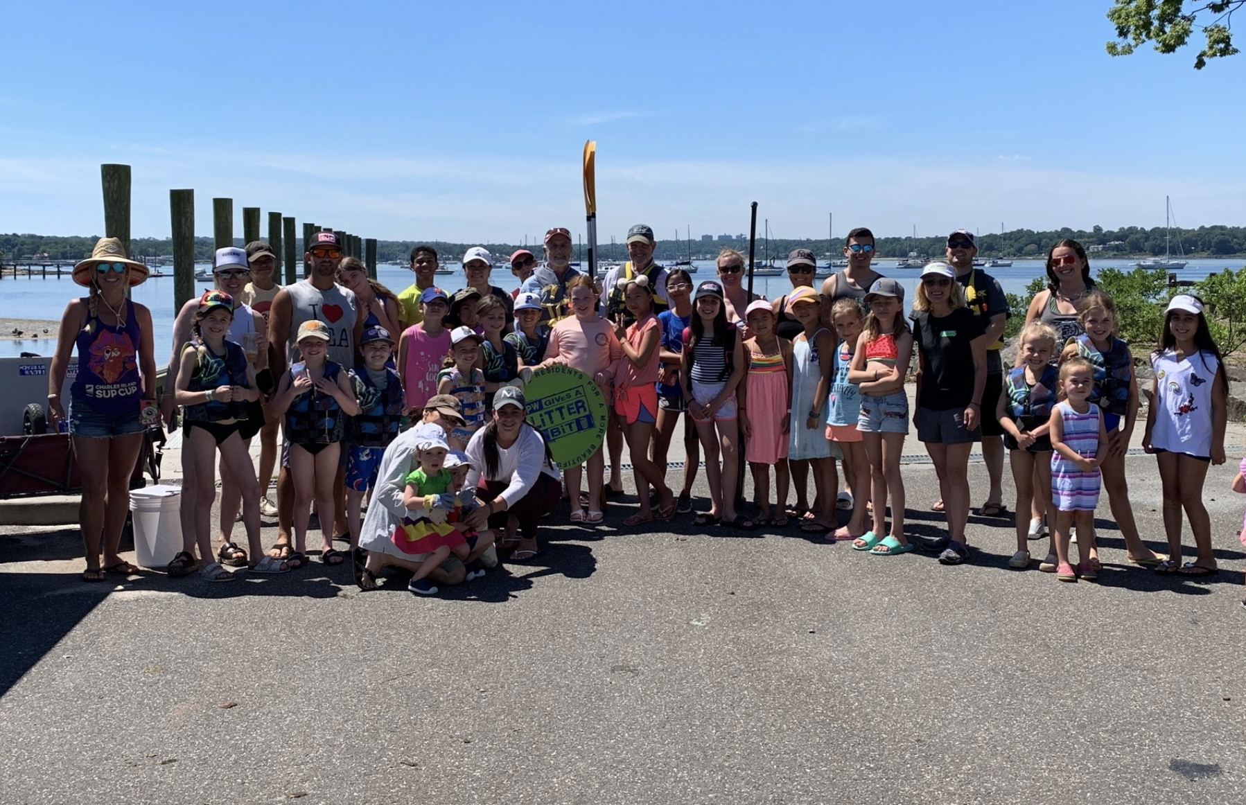 Group of children and adults gathered outdoors near the water, holding a green sign with text, with boats and a dock in the background on a sunny day.