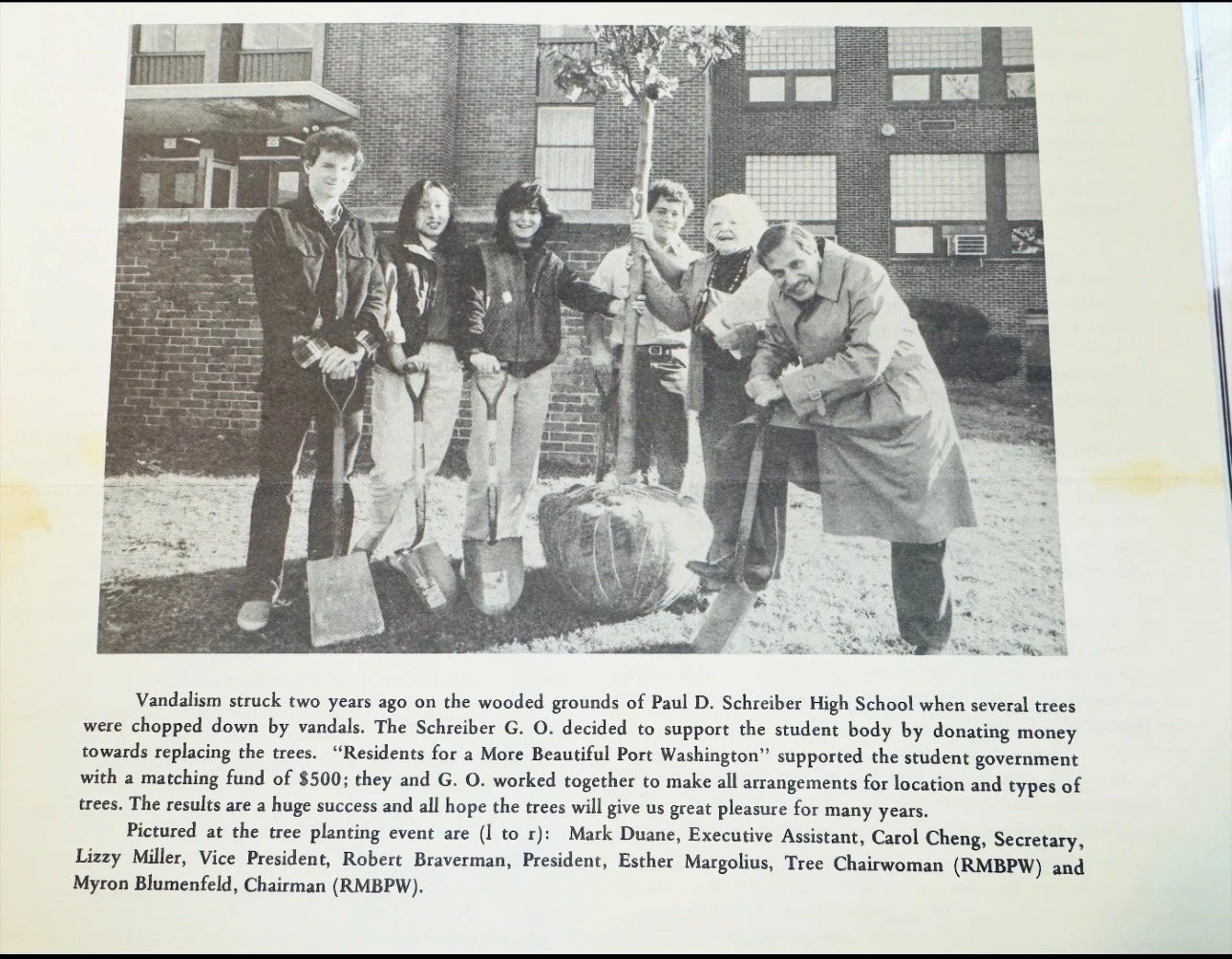 Black and white photo of a group planting a tree in a schoolyard, with a brick building in the background. Seven people hold shovels and a young tree, including children and adults.