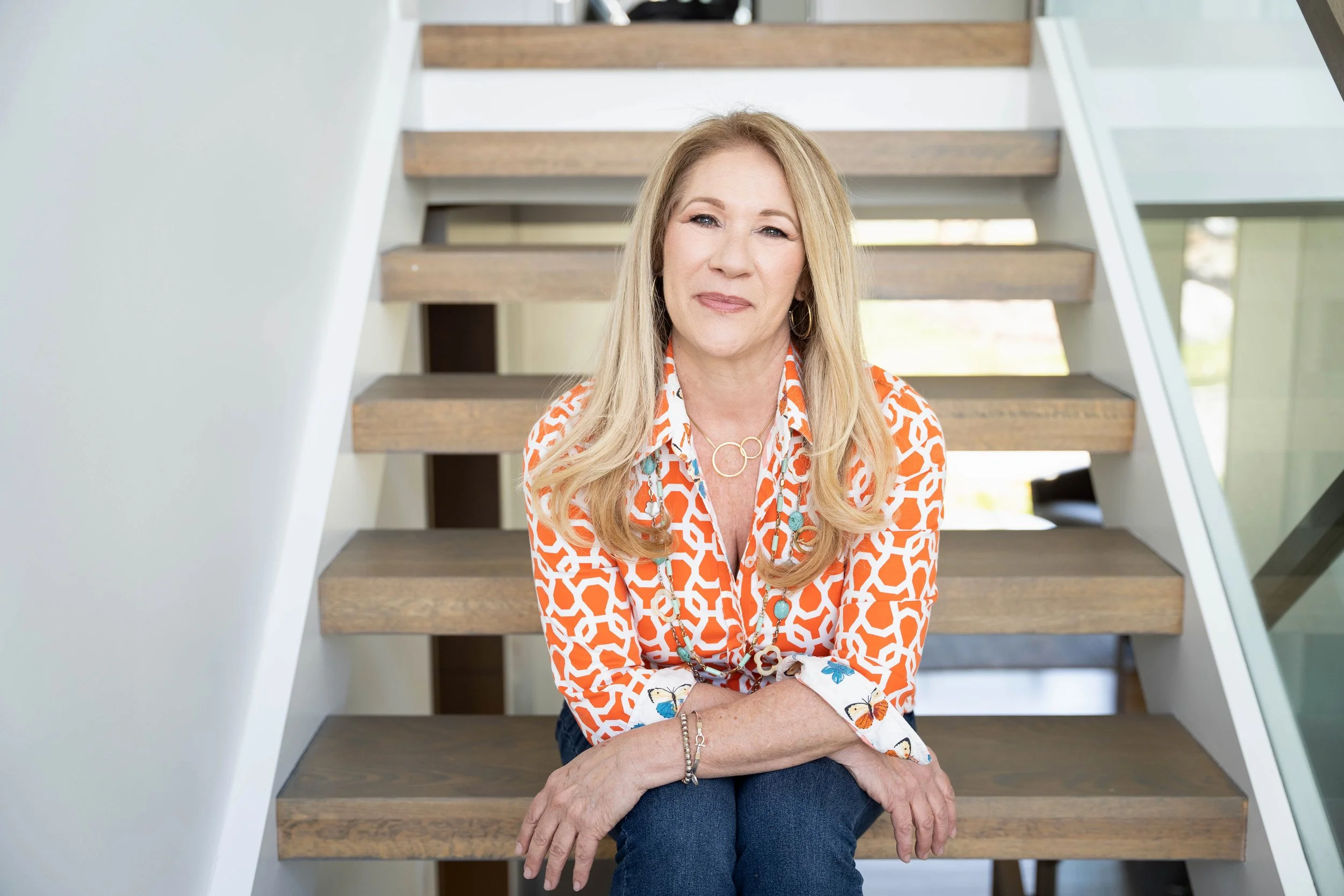 A middle-aged woman with long blonde hair sitting on wooden stairs inside a modern home, wearing an orange and white geometric patterned blouse and a turquoise necklace.