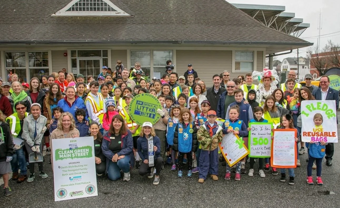A large group of children and adults gathered outside a building for a community cleanup event. Many people are holding signs promoting recycling and reducing waste, and some are wearing reflective vests.