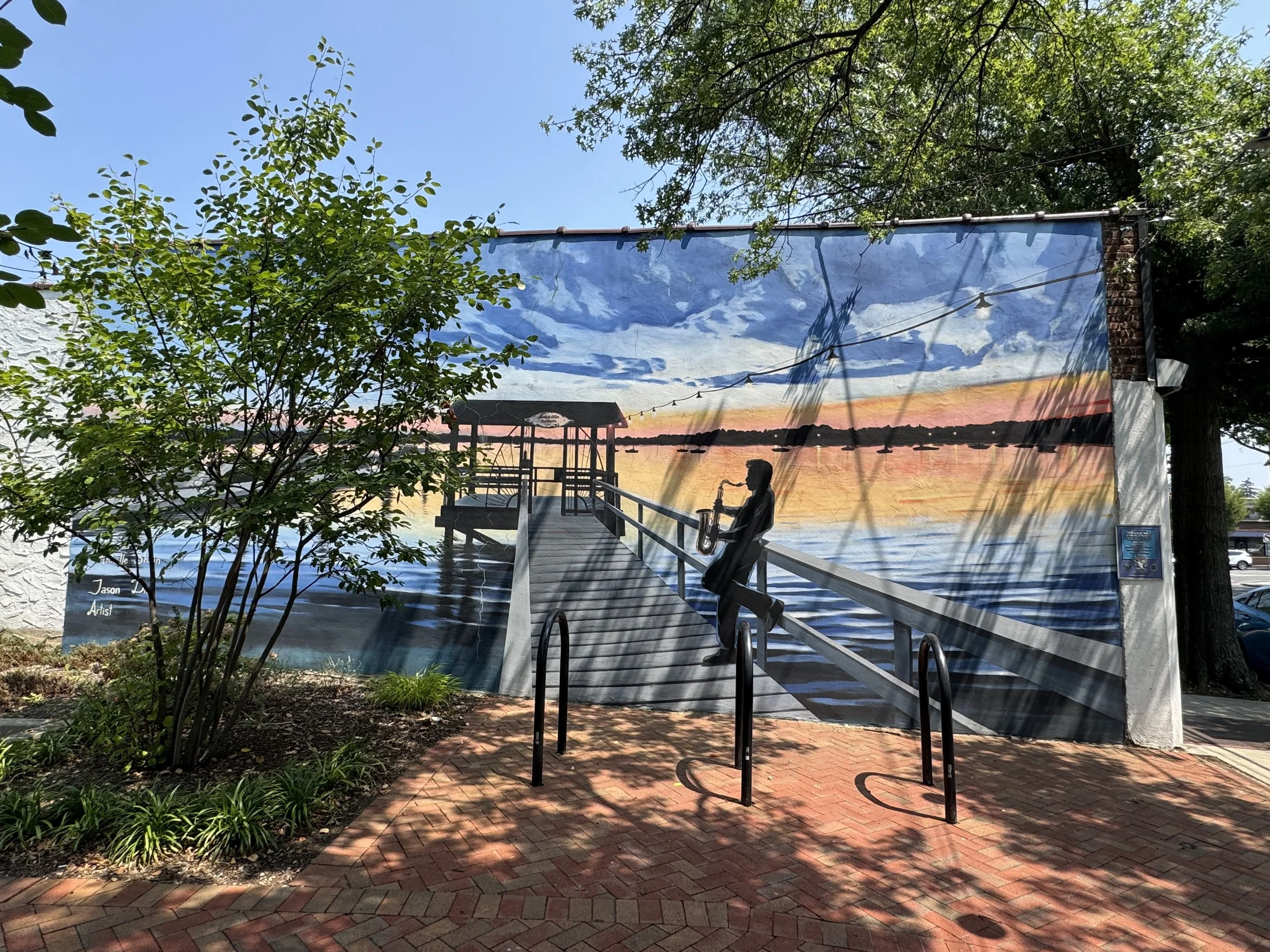 Large mural of a sunset over a lake with a dock, a woman with a saxophone, and boats in the water, painted on an outdoor wall.