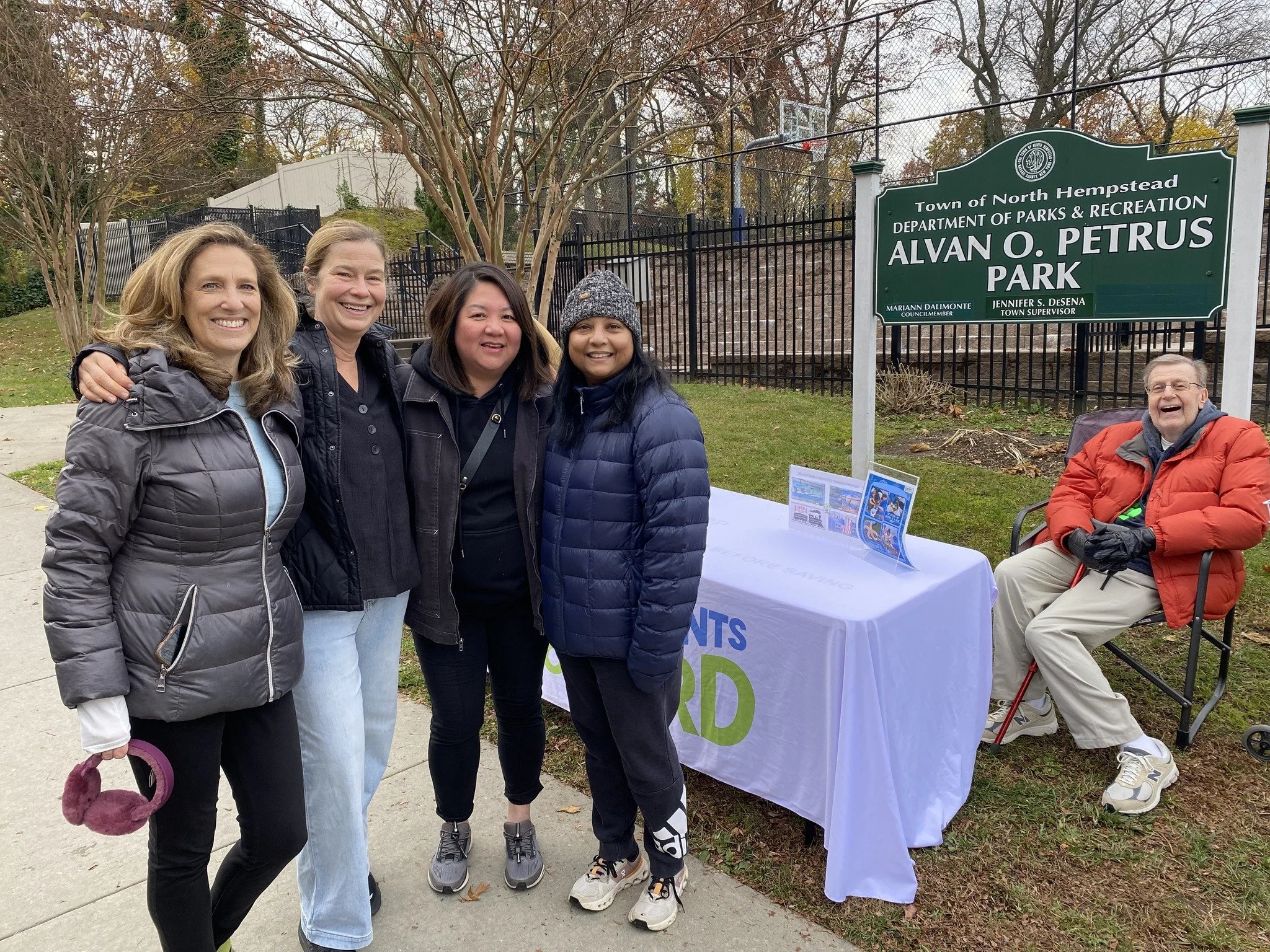 Four women and one man are standing at a park entrance, smiling for the camera. The man is seated near a table with flyers, next to a sign that reads 'Alvan O. Petrus Park' in North Hempstead. The women are wearing jackets and appear to be enjoying a