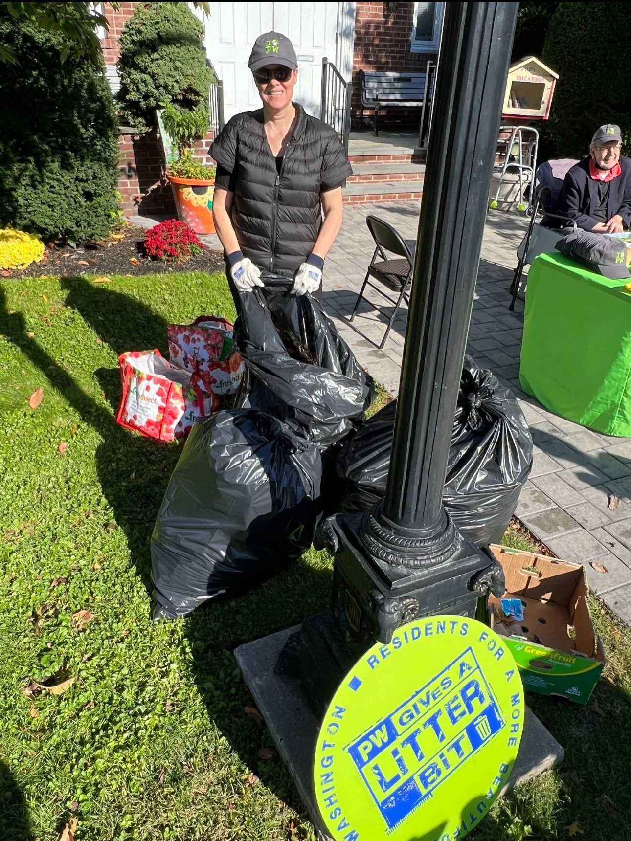 Woman smiling and standing next to plastic bags filled with trash during outdoor litter cleanup event. She is wearing gloves, sunglasses, a gray cap, and a black vest. Behind her is a building with a white door, some potted plants, and volunteers sit