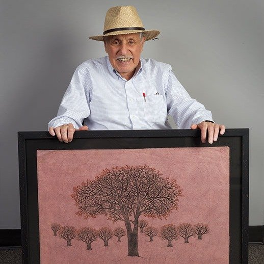 Myron Blumenfeld, the founder of Residents Forward wearing a straw hat and white shirt holding a framed artwork of a tree with many smaller trees below it against a plain gray background.