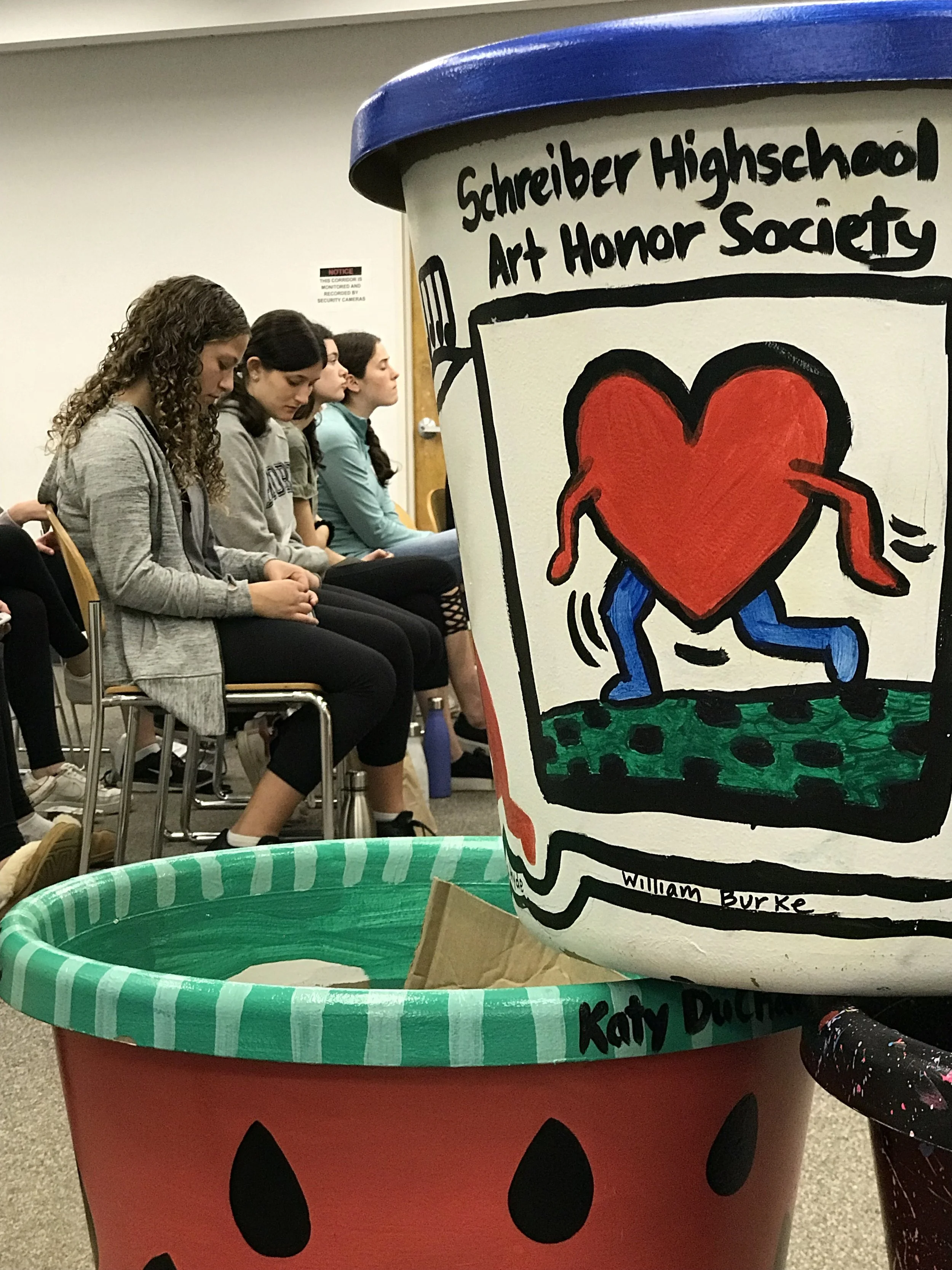 Group of students sitting in chairs during a meeting or presentation, with a large decorated container in the foreground displaying artwork related to the Schreiber High School Art Honor Society.