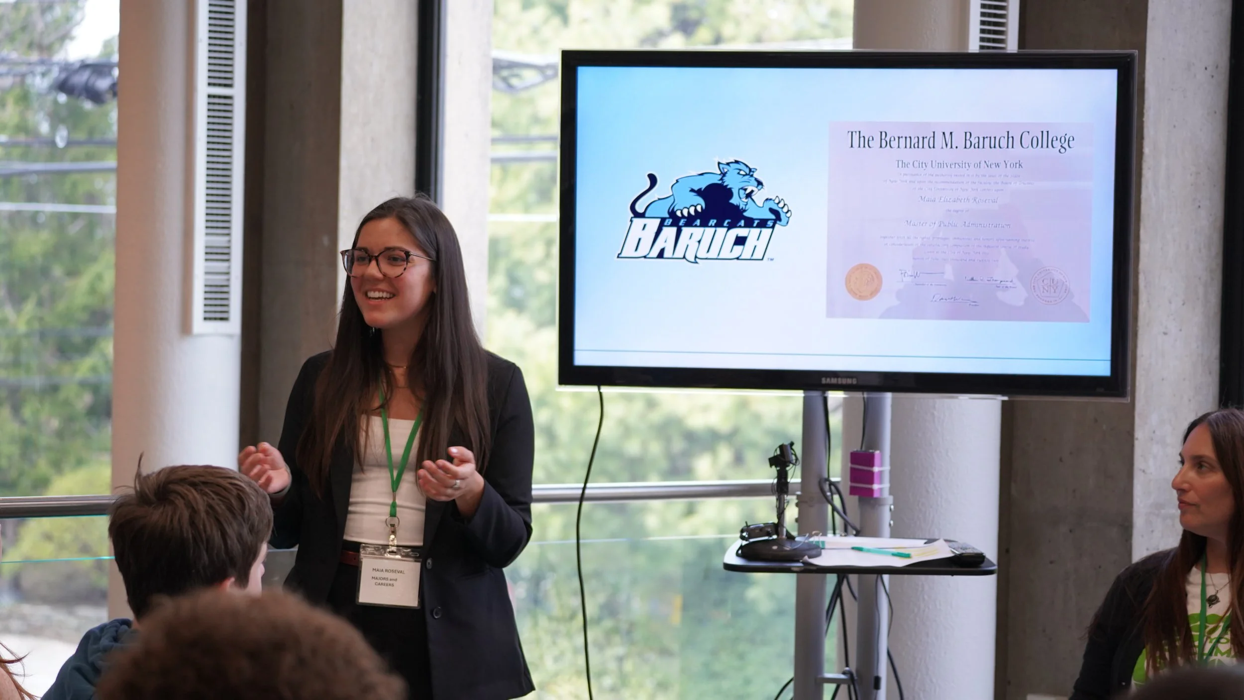 A woman with long brown hair and glasses giving a presentation in a conference room, with a large screen displaying a logo of a blue bearcat for Berkeley College and a diploma from The Bernard M. Baruch College in the background.