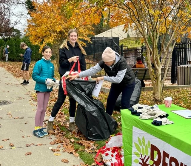Three women and two children participating in a neighborhood cleanup, collecting leaves and trash, with a table of supplies and an autumn tree in the background.
