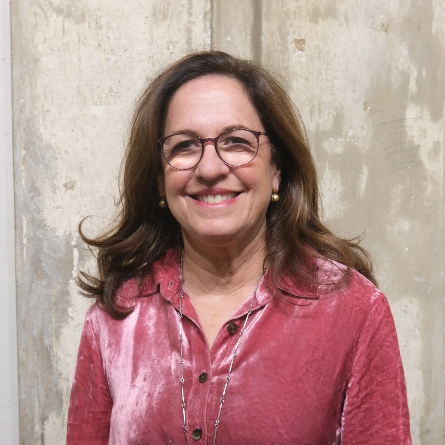 Smiling woman with glasses and brown hair, wearing a pink velvet blouse, standing in front of a textured beige wall.