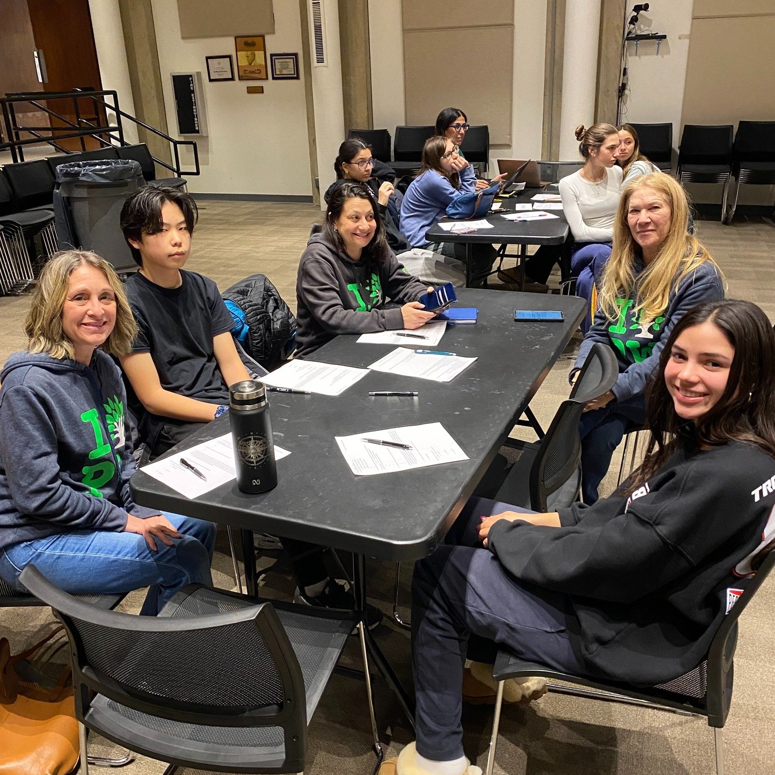 Group of seven people sitting at a rectangular table in a room with beige walls. They have papers, pens, and coffee cups, appearing to be in a meeting or workshop. Some are smiling, and some are looking at their devices or papers.