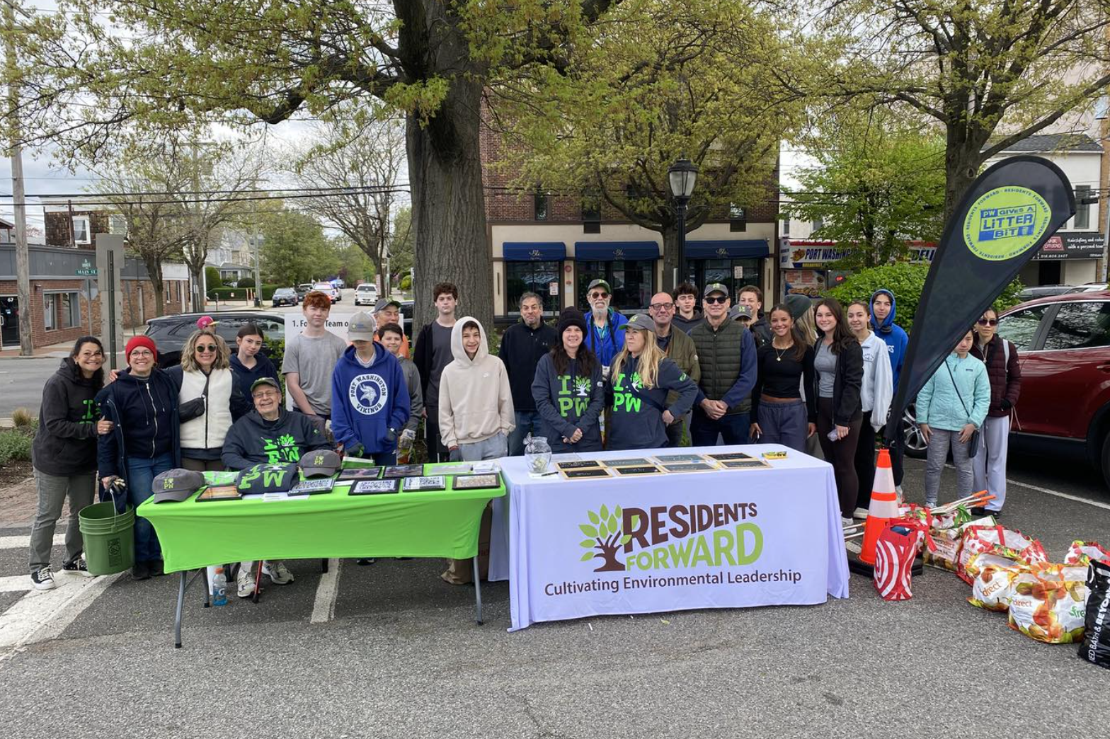 Group of people gathered outdoors for an environmental event, some wearing Resident Forward shirts, with tables displaying materials and supplies, and recycling or waste bags on the ground.