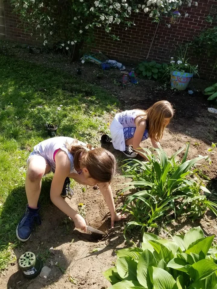 Two young girls planting or gardening in a backyard garden with a brick wall, flowers, and green plants around them.