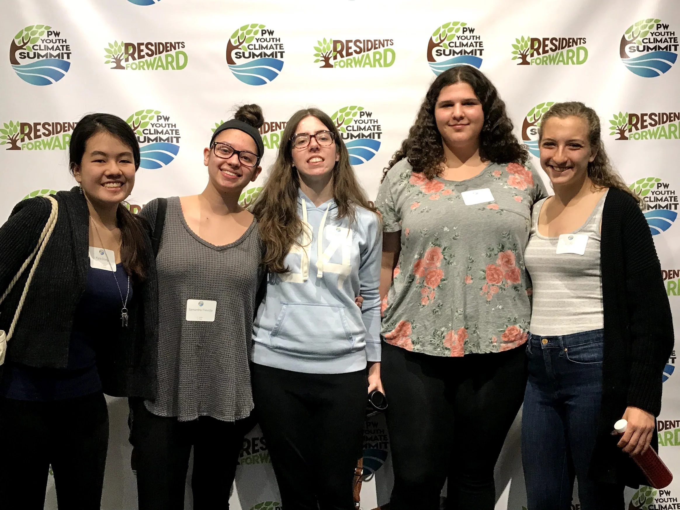 Group of five young women standing together in front of a backdrop with logos for the PW Youth Climate Summit and Residents Forward, smiling for the photo.