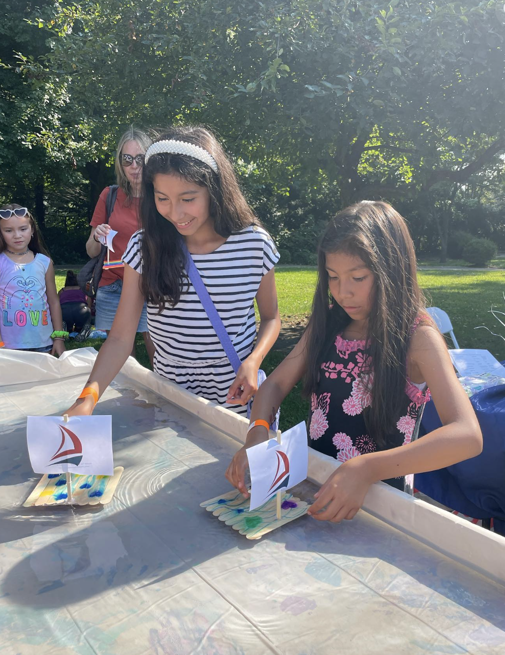 Two young girls are playing with small sailboat crafts on a water table outdoors, with smiling faces and enjoying a sunny day. An adult woman and another girl are visible in the background, standing near green trees and grass.