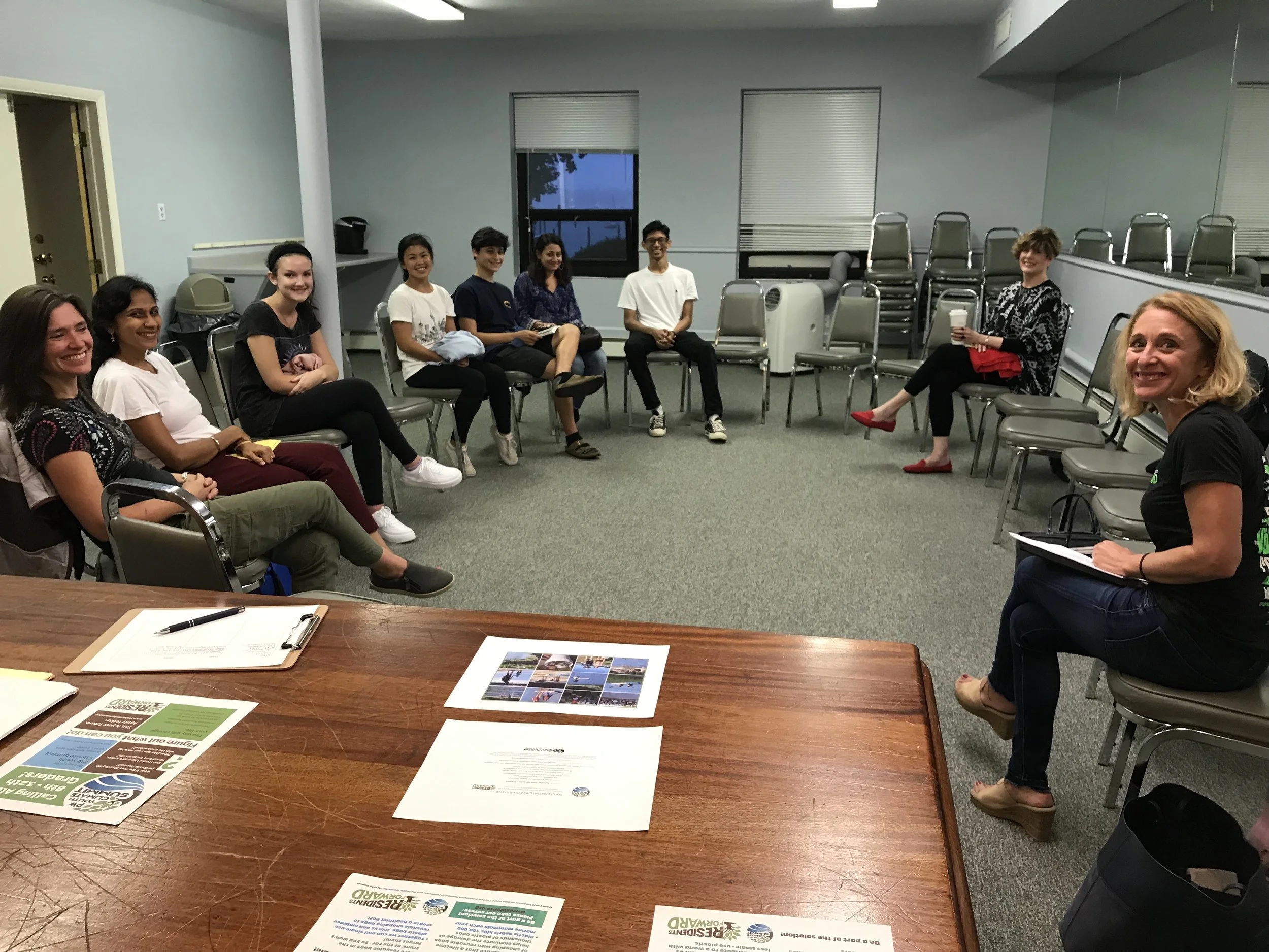 A group of people sitting in a circle in a meeting room, smiling and engaging, with a woman at the front taking notes and various papers and pamphlets on a table in the foreground.
