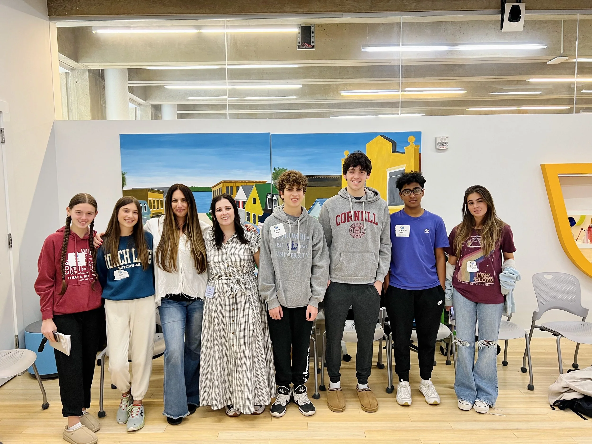 A group of nine young people standing in a row indoors with a colorful mural of houses and a river behind them. They are smiling and wearing casual clothing, some with name tags.