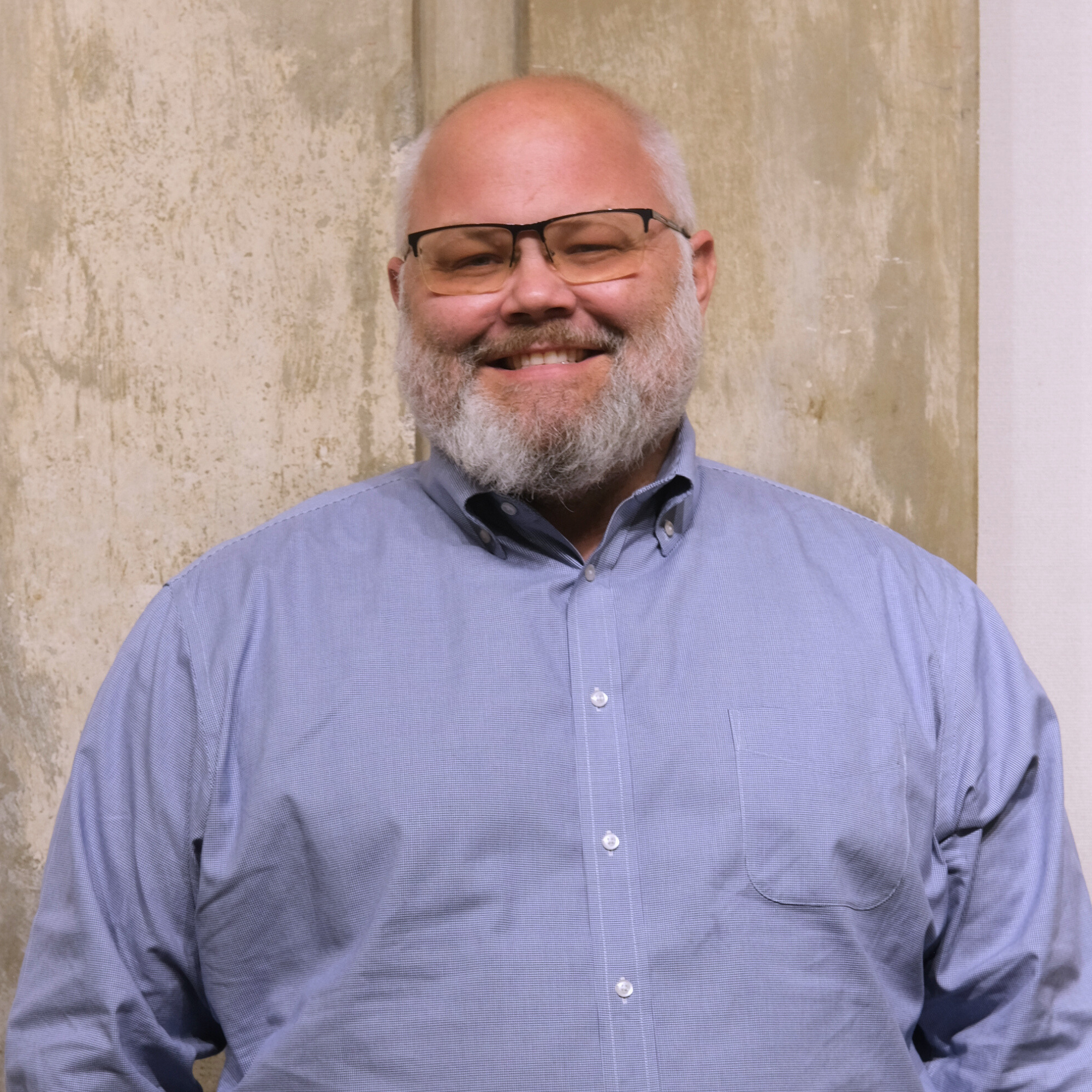A smiling man with glasses and a beard wearing a light blue collared shirt standing in front of a textured beige wall.