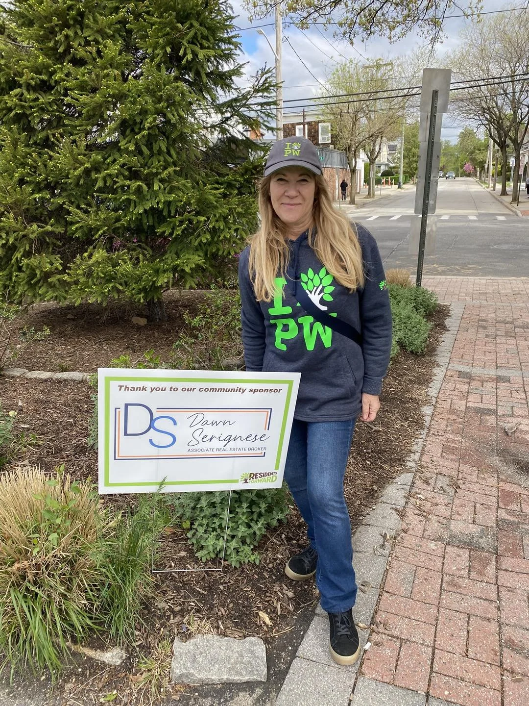 A woman standing on a sidewalk next to a tree, wearing a dark hoodie and cap with green lettering, holding a sign thanking a community sponsor for supporting residents forward.