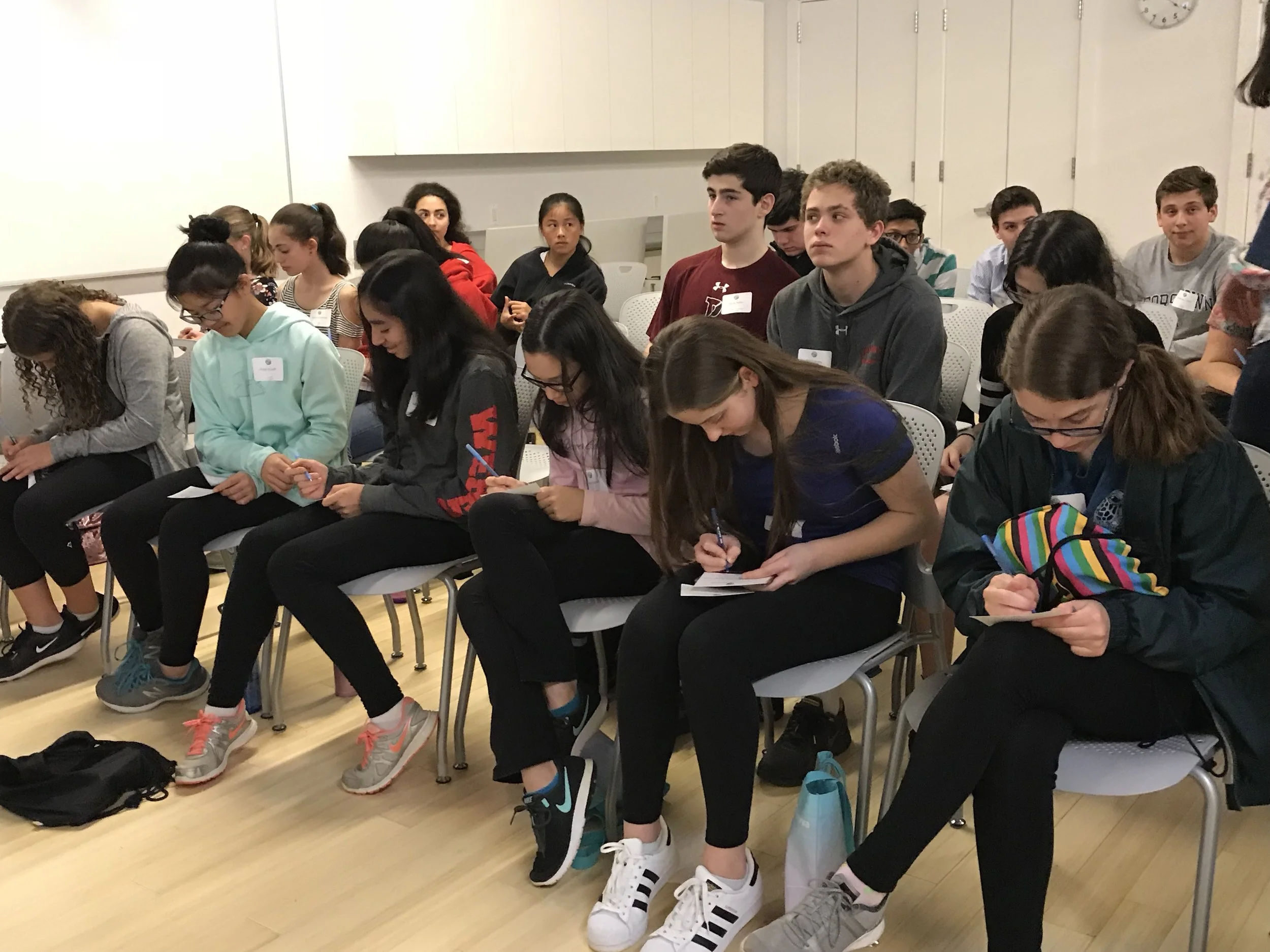 Group of teenagers seated in a classroom, some taking notes and others listening attentively.