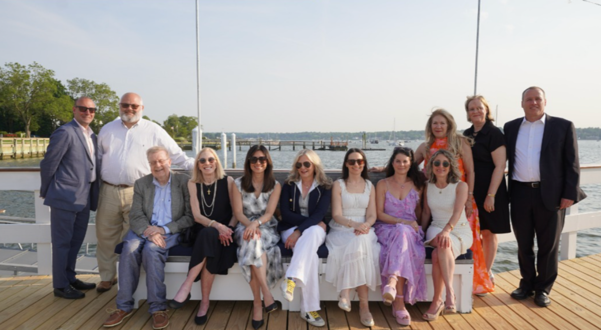 Group of 12 people, both men and women, posing for a photo on a boat dock near water, dressed in casual and semi-formal clothing, with a harbor and boats in the background.