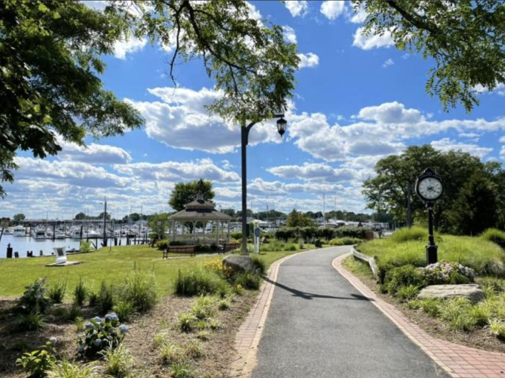 A park pathway leading to a gazebo near a marina with boats, under a partly cloudy sky, surrounded by greenery and flowers, with a clock on a pole and street lamps.