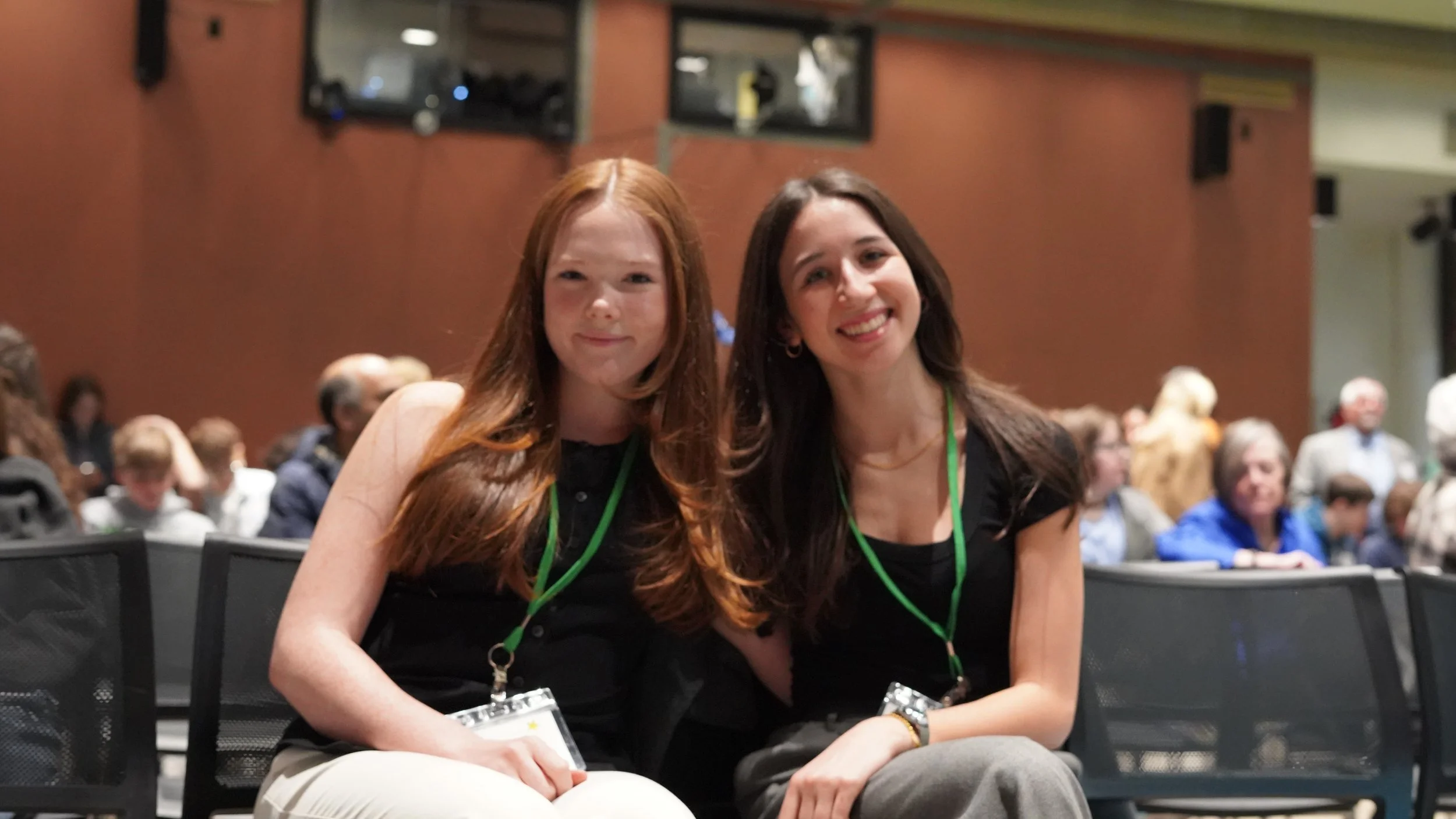 Two young women sitting together at a conference or event, smiling at the camera, wearing black tops and green lanyards, with a crowd of people in the background.