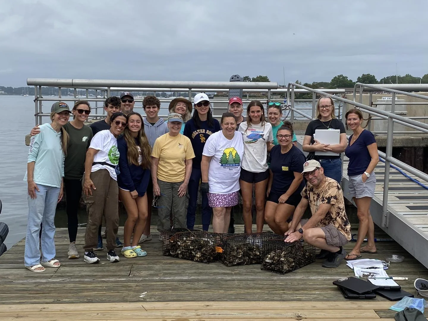 Group of people on a dock, smiling, with cages of oysters in front of them, holding tools and papers, overcast sky, waterfront background.