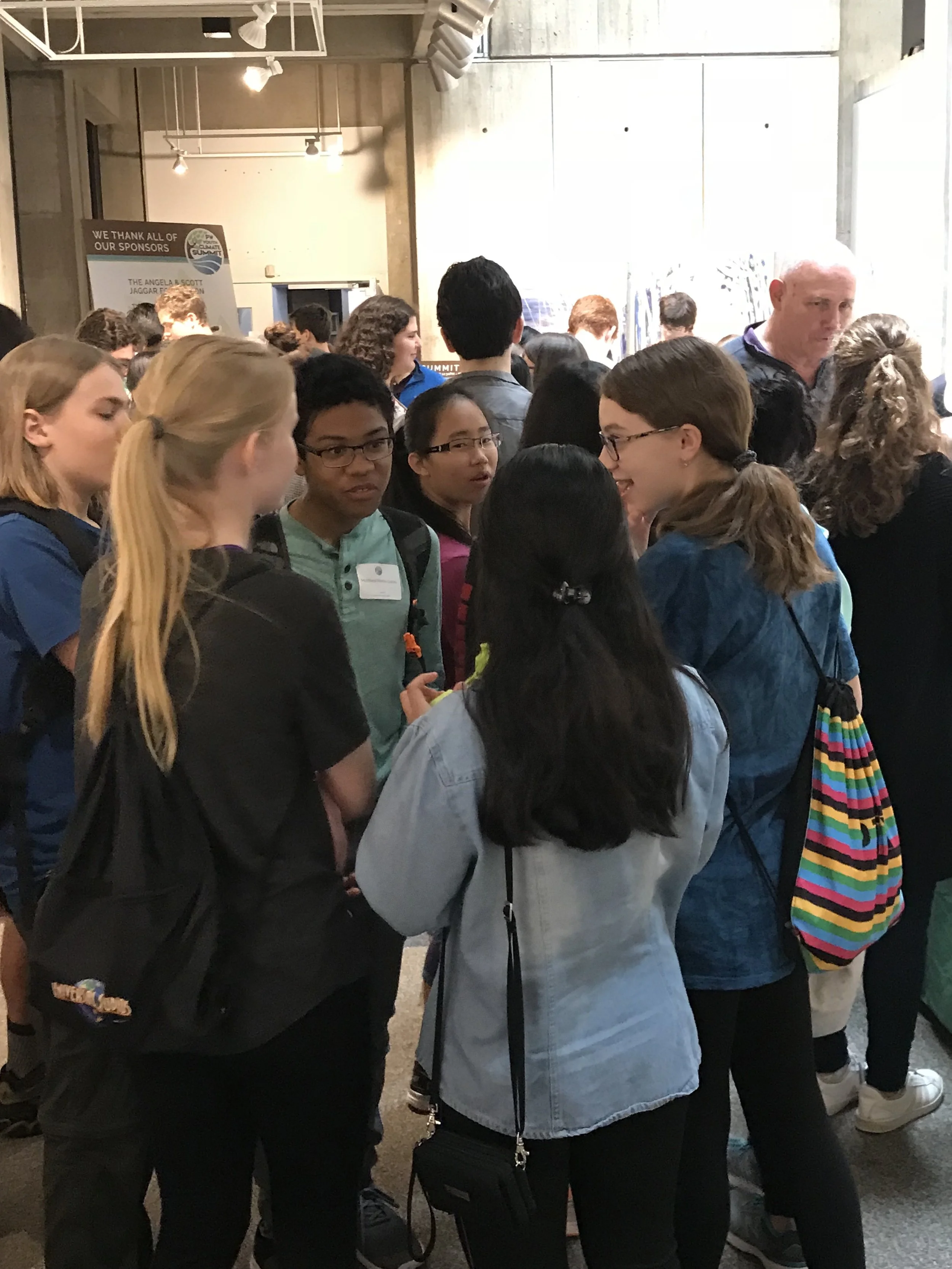 Group of teenagers and adults at an indoor event, engaged in conversation and looking at displays or artwork.