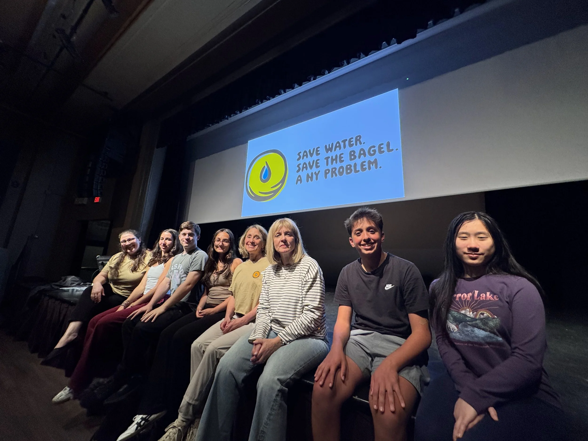 Group of eight people sitting on a stage before a large screen that displays a message promoting water conservation, with the text 'Save water. Save the bagel. A my problem' and a logo of a water drop.