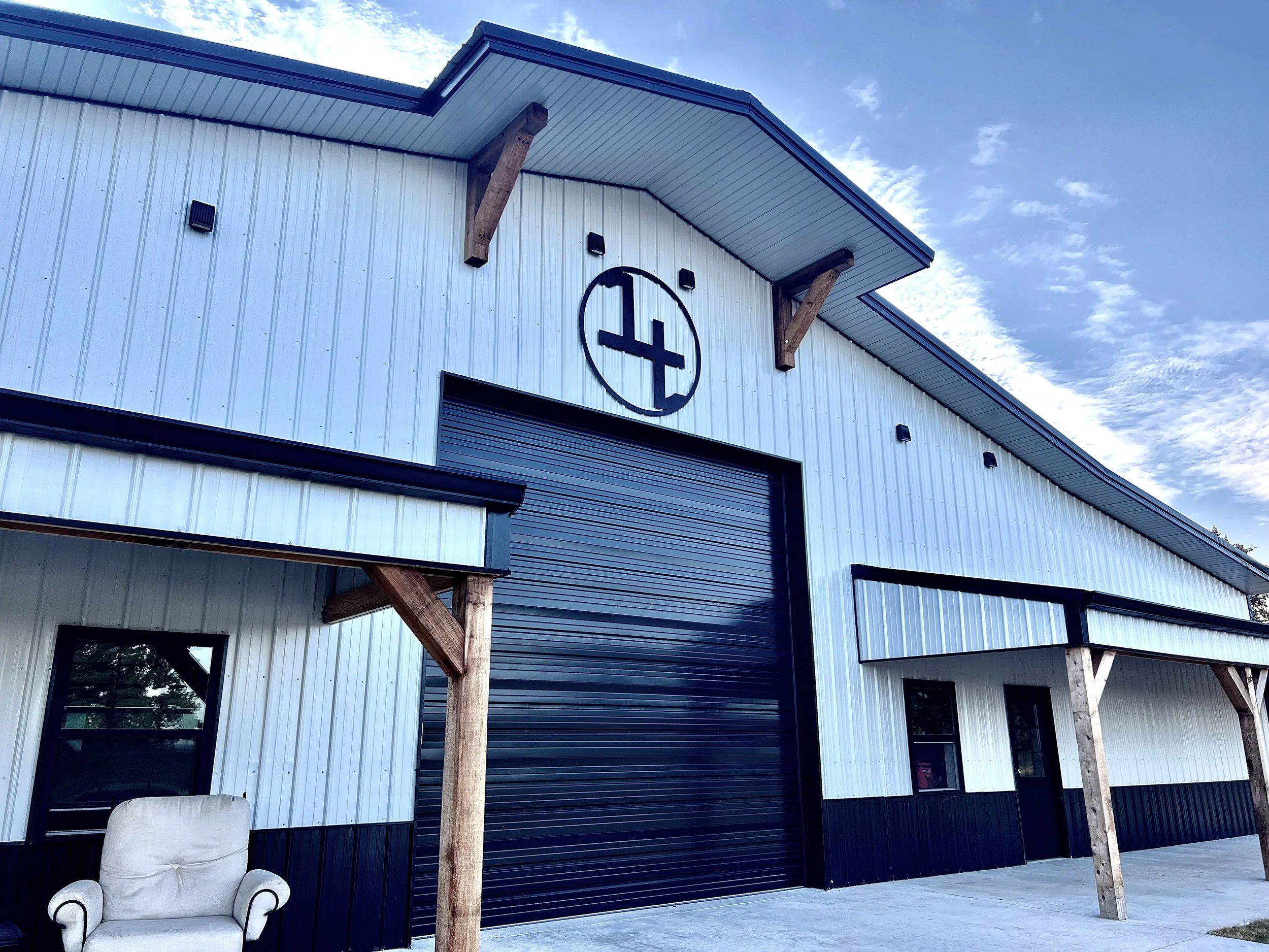 Exterior of a white metal building with wooden posts, a large black garage door, and a logo above the door. A beige armchair sits under an overhang.